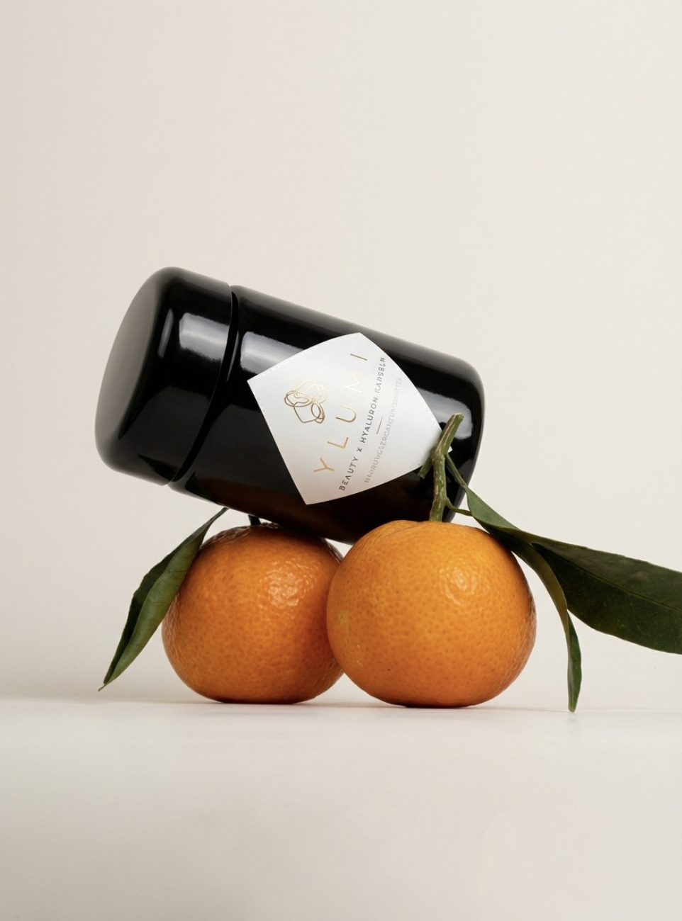 A black jar labeled 'YLU Beauty + Hyaluronic Acid' resting on two oranges with green leaves against a plain white background.