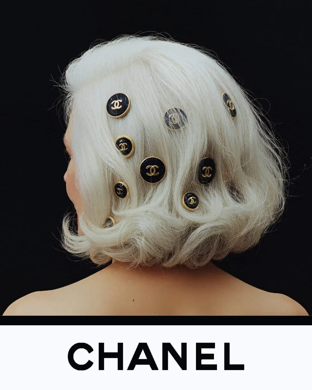 Back view of a person with platinum blonde hair decorated with black and gold Chanel buttons, with the word 'CHANEL' at the bottom.