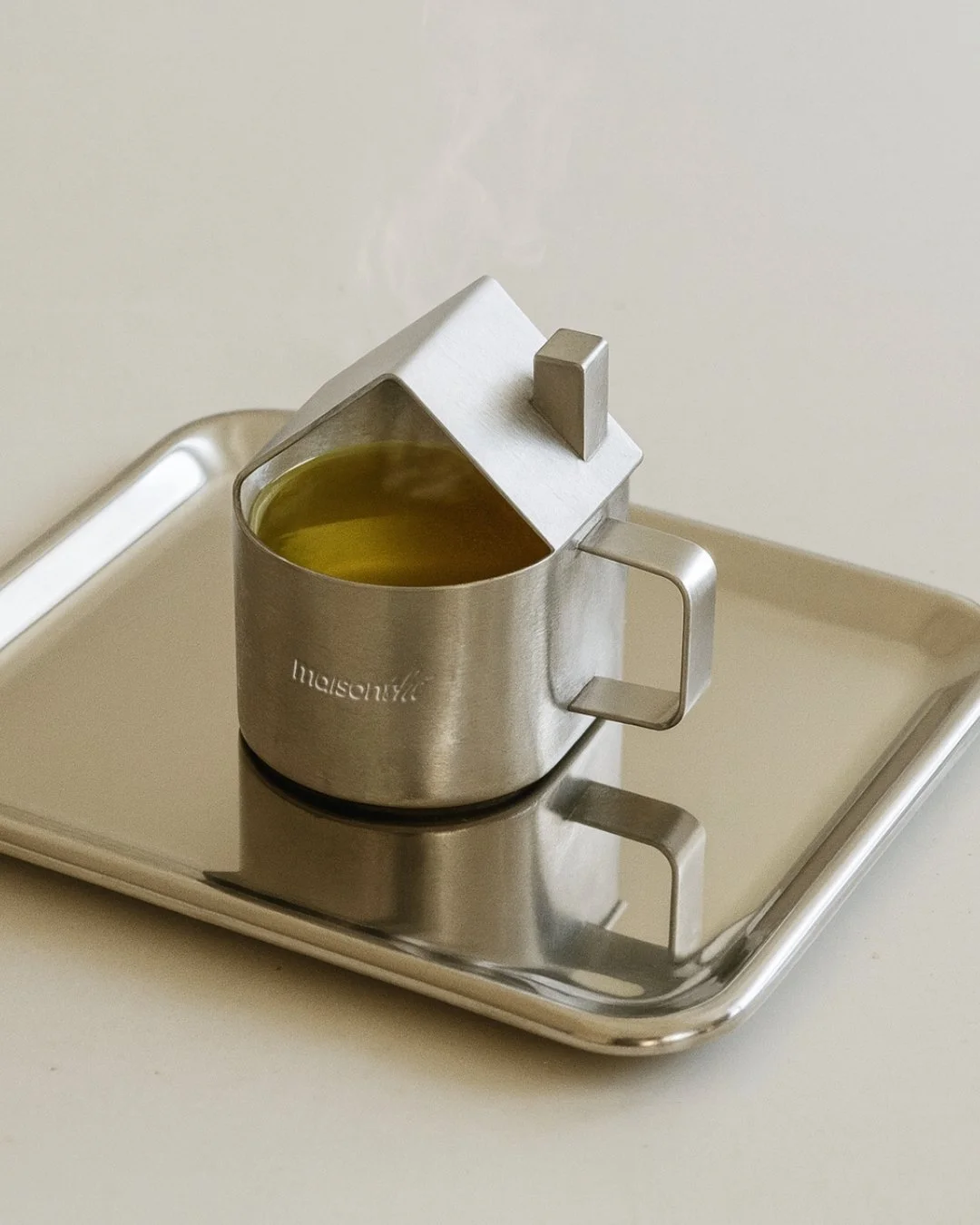 A stainless steel pitcher with a house-shaped lid, filled with green tea, placed on a metal tray.