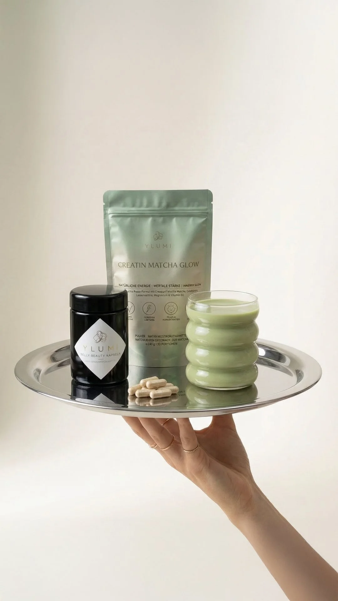 A silver tray held by a hand, containing a black jar, a packet of Y LUMI creatin matcha glow powder, a green ceramic cup with matcha, a few capsules, and a black and white Y LUMI supplement container.