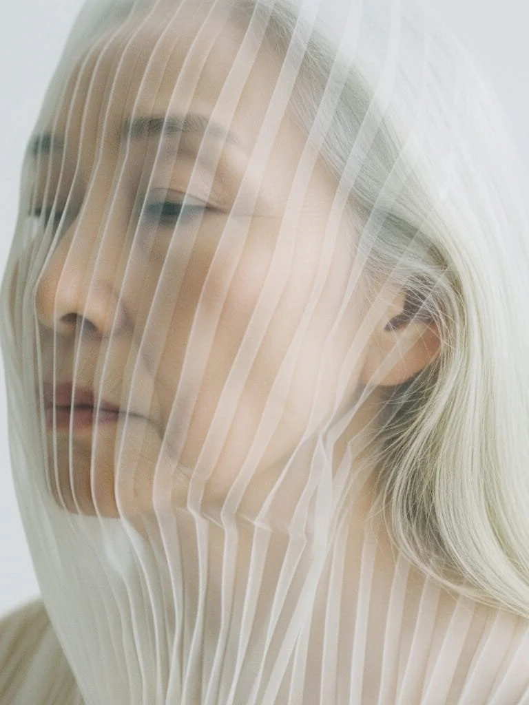 A woman with platinum blonde hair and closed eyes is covered by a translucent, pleated, white fabric that partially obscures her face.