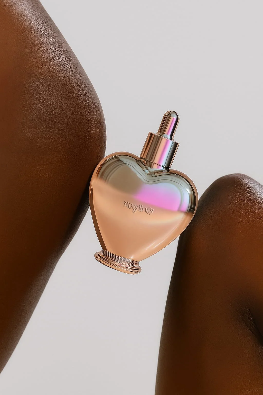 A heart-shaped perfume bottle with iridescent surface resting between dark-skinned thighs against a plain white background.