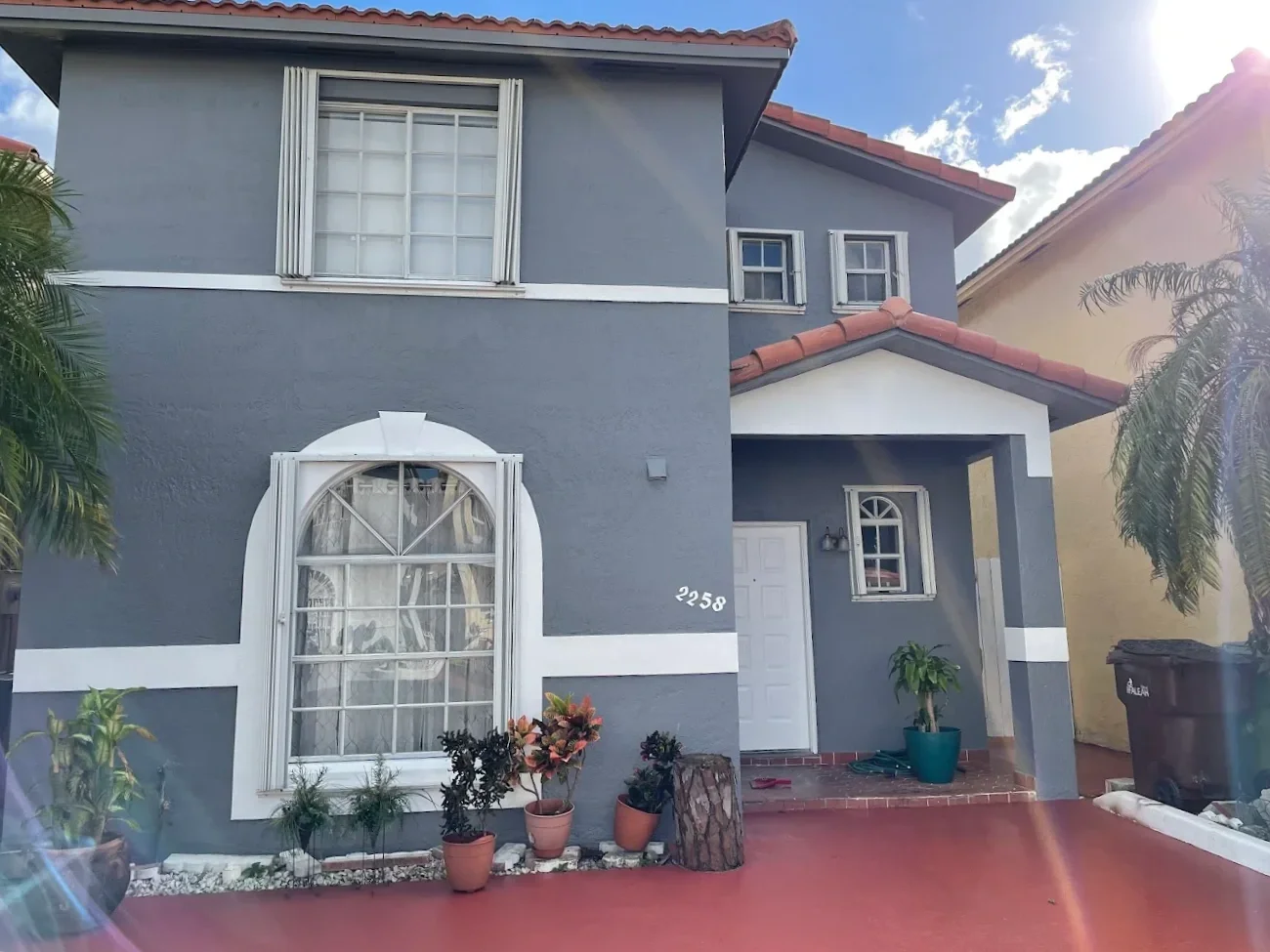 Two-story gray house with white window trims and red-tiled roof, decorated with potted plants in front entrance, under partly cloudy sky.