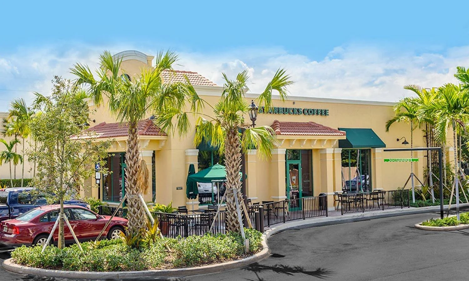 A Starbucks Coffee shop in a tropical setting with palm trees, parked cars, and a clear blue sky.