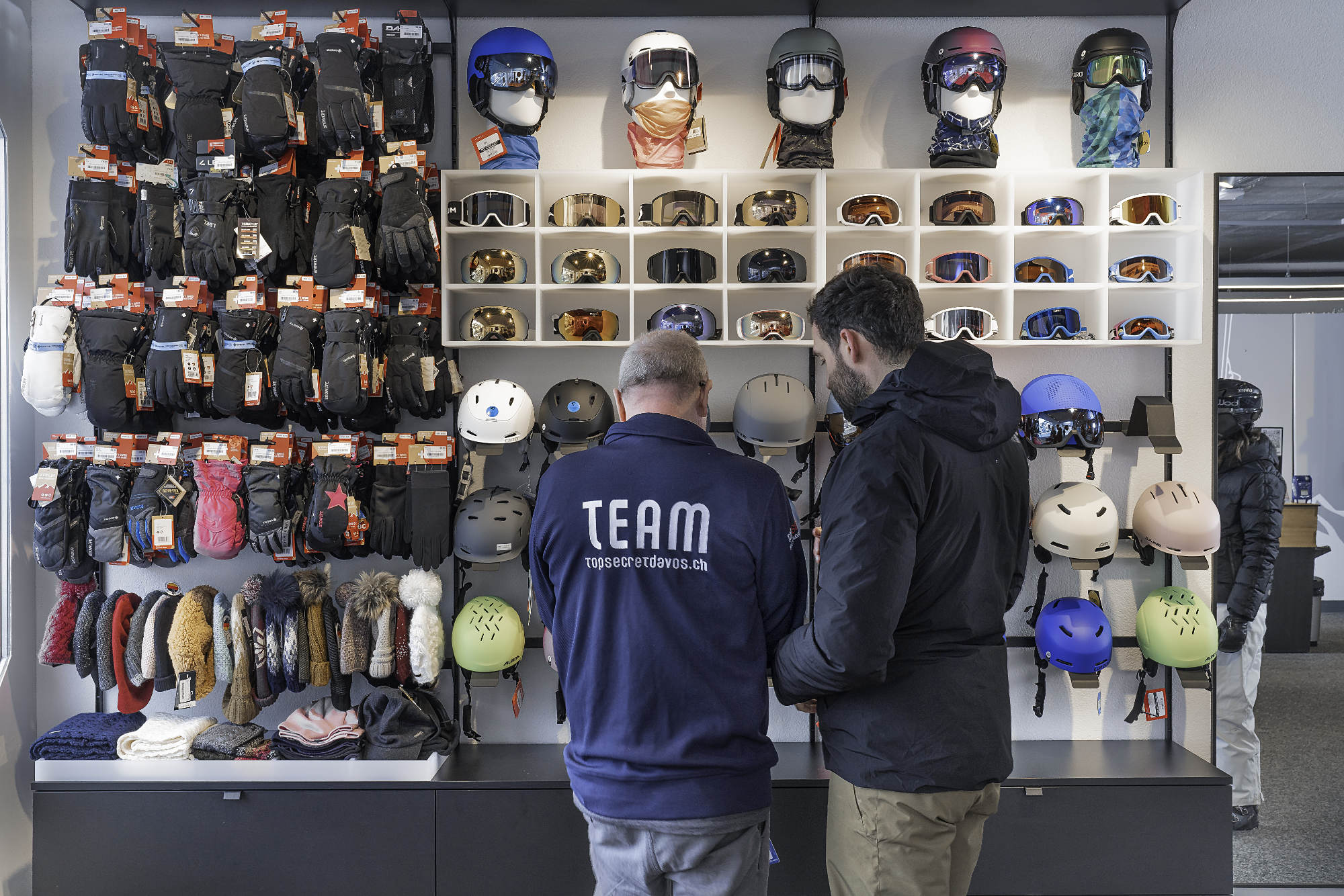 Zwei Männer stehen vor einem Wandregal mit Skibrillen, Helmen, Handschuhen und Mützen in einem Sportgeschäft, wobei sie miteinander sprechen.