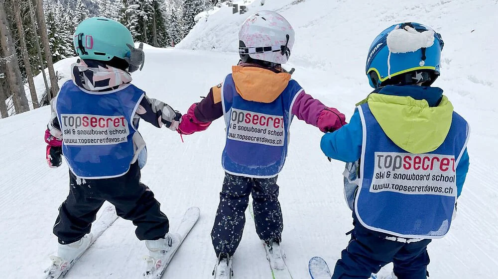 Drei Kinder beim Skifahren auf einer verschneiten Piste, alle tragen Helme und blaue Sicherheitswesten mit dem Logo 'top secret ski & snowboard school'.