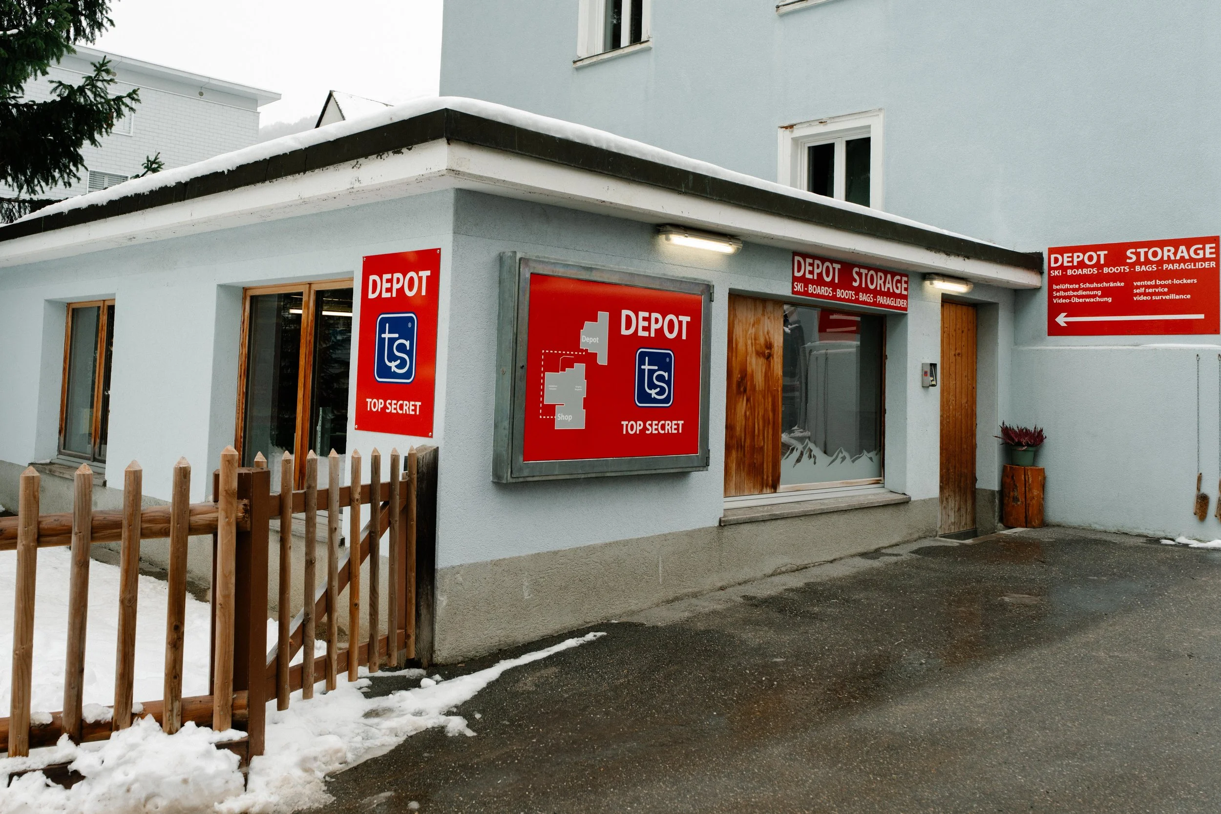 Äußerer Teil eines Gebäudes mit Schildern, die auf Lager für Skier, Boote, Taschen und Paragleiter hinweisen, im Winter mit Schneeregen und Schneespuren auf dem Boden.
