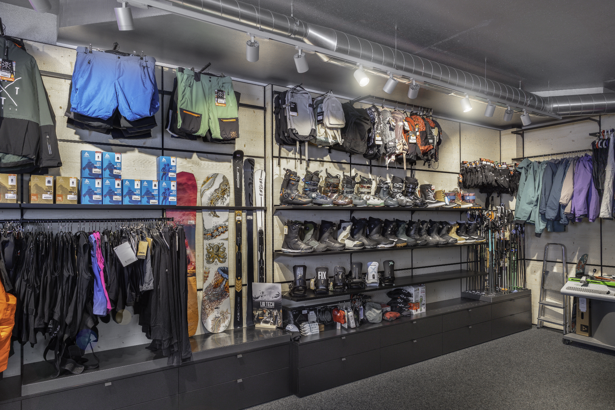 Beschreibung eines Sport- bzw. Outdoor-Ladenregals mit Skiausrüstung, Wanderrucksack, Ski, Skischuhe, Snowboards, Snowboardboots, Accessoires, und Winterkleidung.