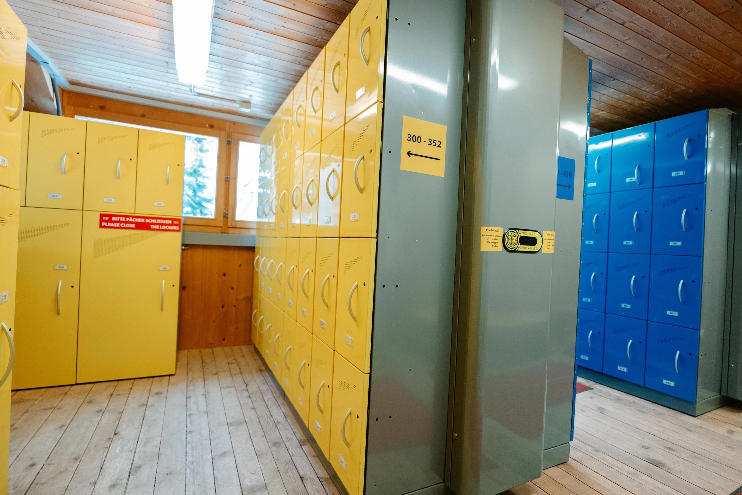 Mehrere gelbe und blaue Schließfächer in einem Flur, mit Holzdecken und Holzboden, Beschriftung auf einigen Schließfächern.