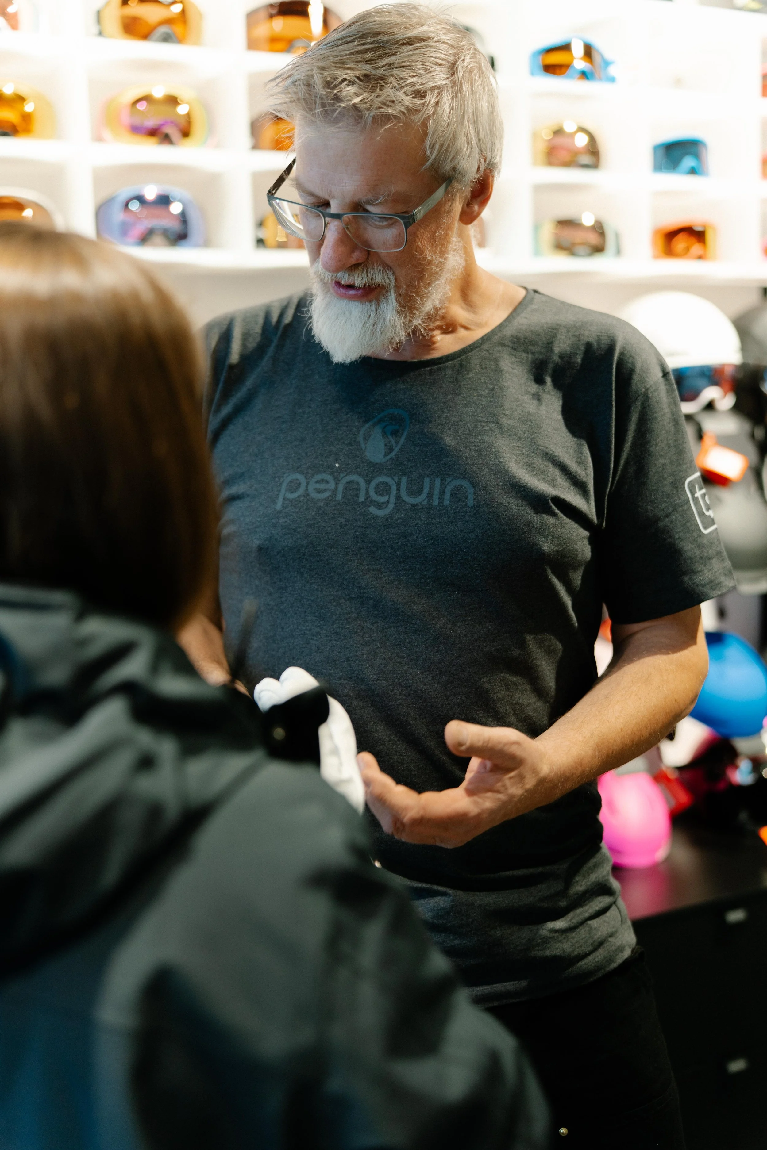 Ein älterer Mann mit grauem Bart und Gläsern trägt ein dunkelgraues T-Shirt mit dem Logo 'penguin' und spricht mit einer Person. Im Hintergrund sind helle Wandregale mit verzierten Skibrillen und Helmen.