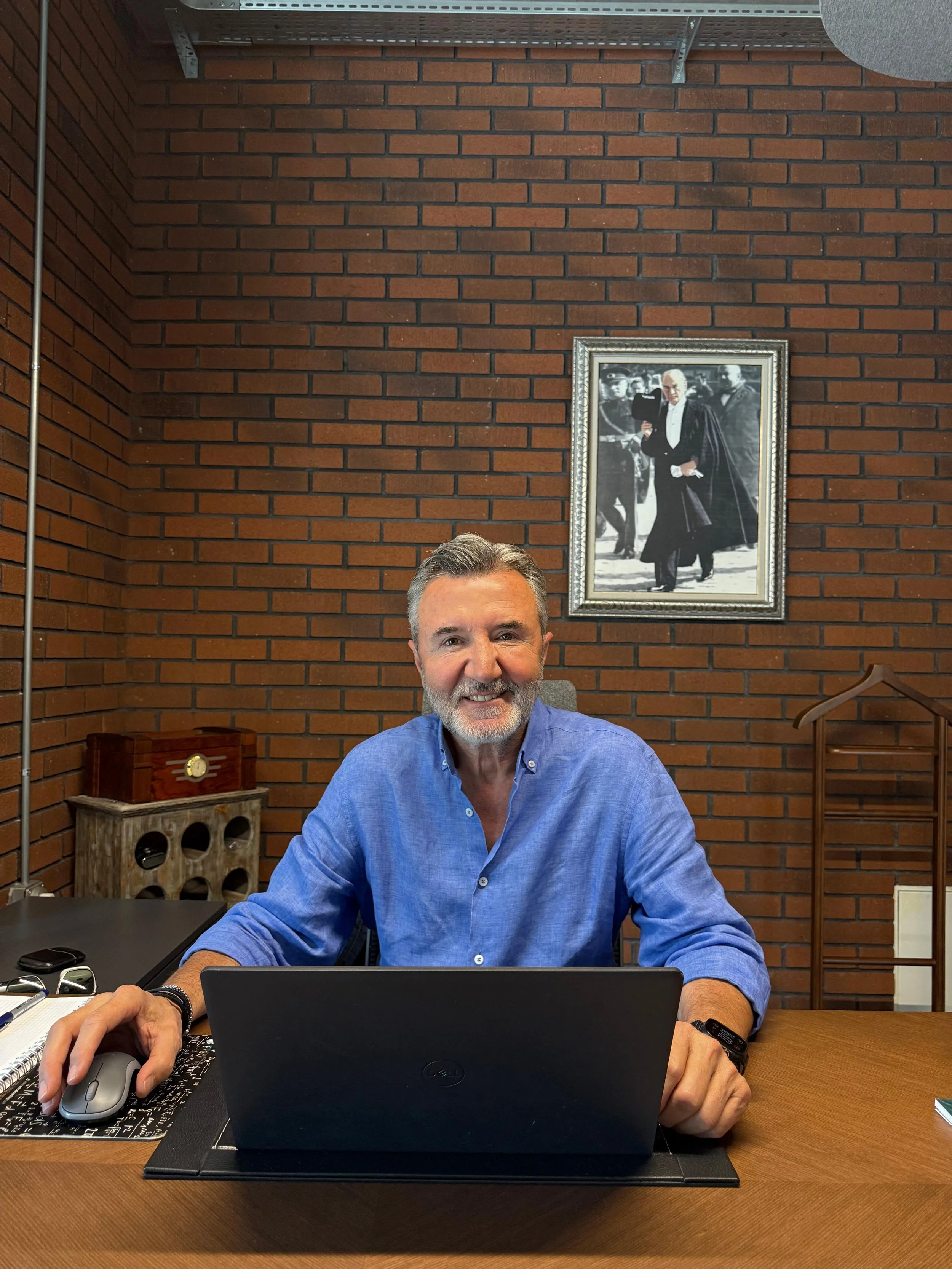 A man with gray hair and a beard, wearing a blue button-up shirt, sitting at a desk with a laptop, mouse, and notepad. The background features a brick wall, a framed black-and-white photo of a man in a cape, and some furniture.