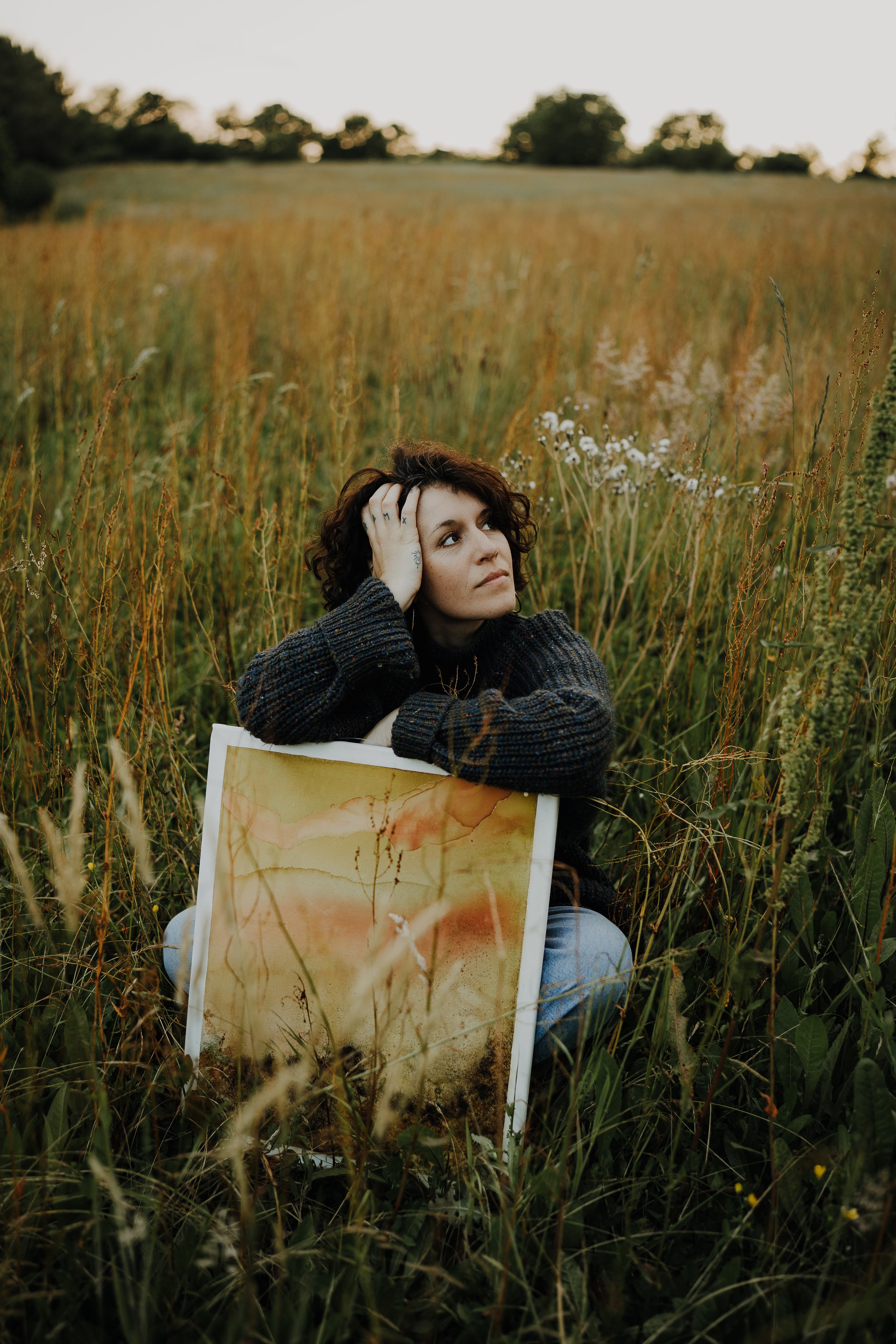 Une jeune femme aux cheveux bouclés bruns, portant un pull noir, assise dans une prairie avec de longues herbes et des fleurs blanches. Elle pose sa tête sur sa main gauche, regardant vers le haut. Elle tient une œuvre d'art ou une affiche avec des couleurs chaudes en arrière-plan dans ses genoux.