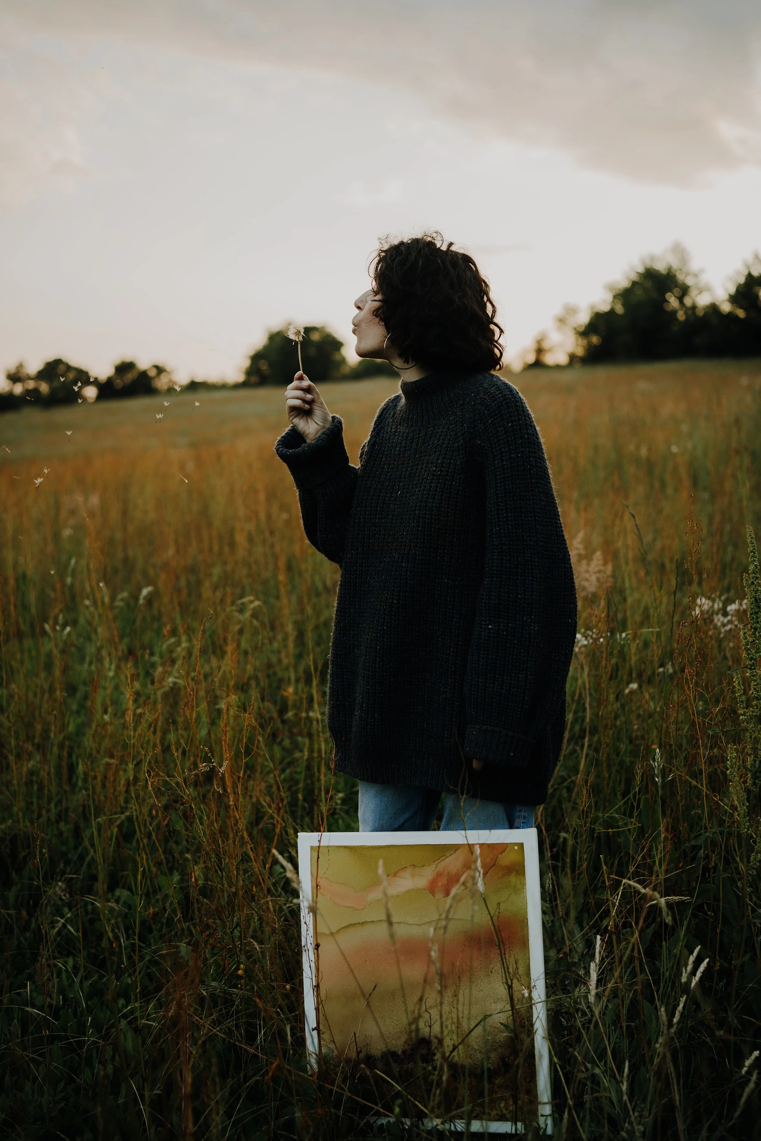 Une femme avec des cheveux bouclés portant un pull noir se tient dans un champ pendant le coucher du soleil, soufflant des graines de dandelion.
