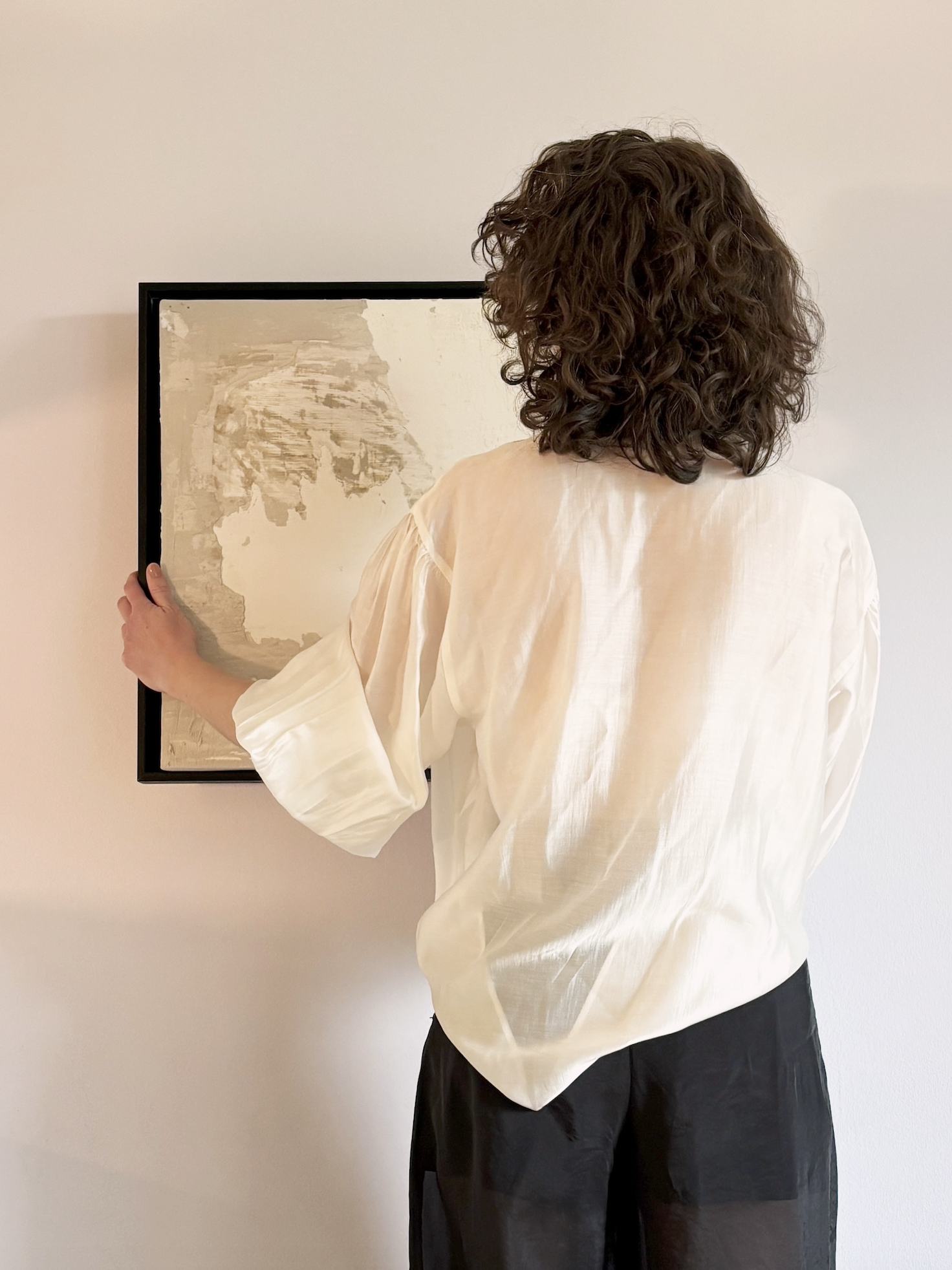 Eine Person mit lockigem braunem Haar, die ein beige Hemd trägt, hält ein Kunstwerk in einem schwarzen Rahmen vor einer weißen Wand.