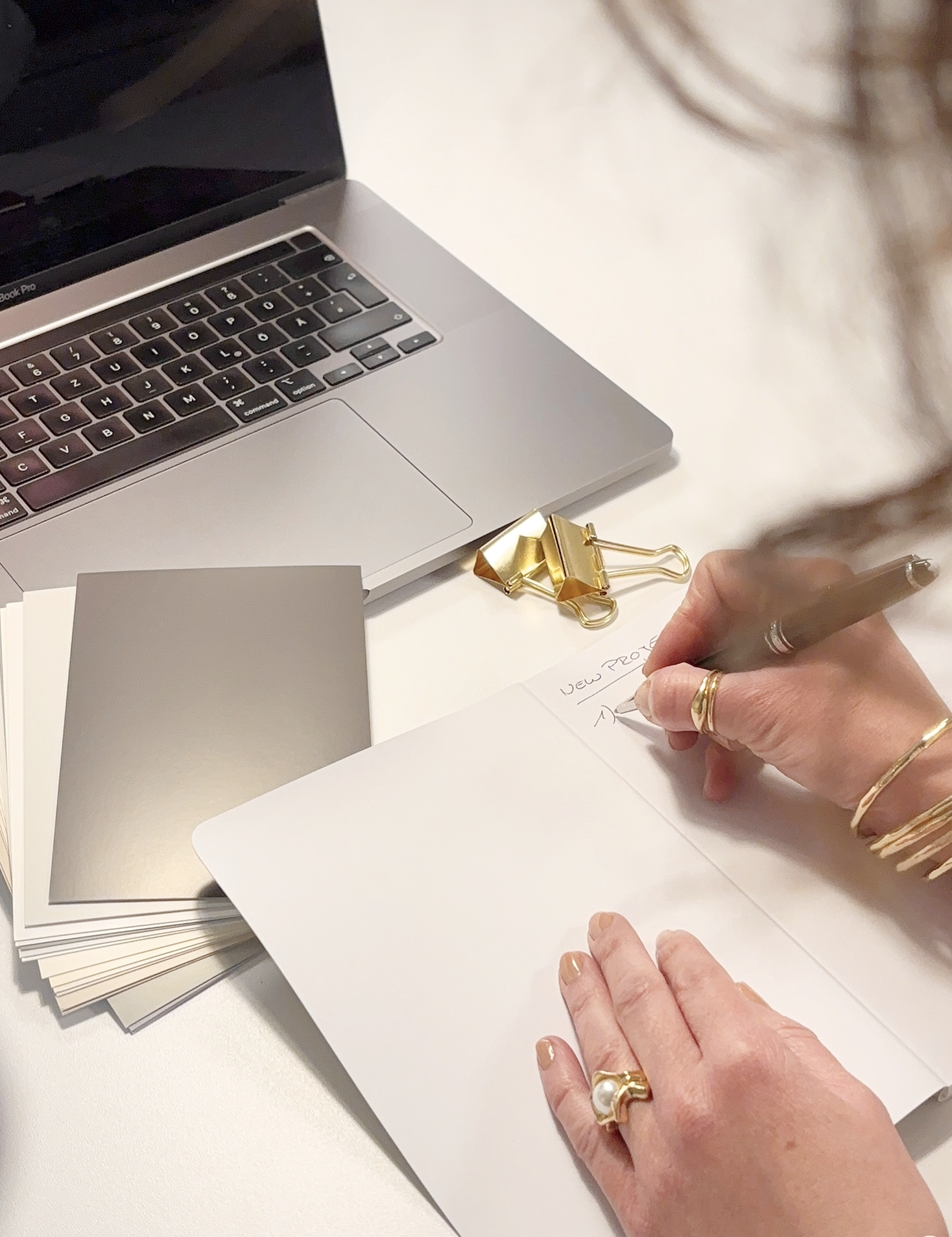 Hand einer Person schreibt in ein Notizbuch, umgeben von einem Laptop, Notizkarten, einer goldenen Klammer und einem Goldring. Es scheint, dass die Person eine To-Do-Liste oder Notizen macht.
