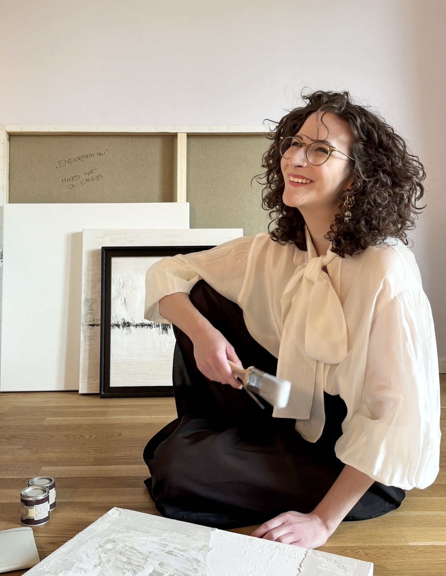 Frau mit Locken, Saskia Bartels, Brille und Earring, sitzt im Kunststudio, malt auf Leinwand und lächelt.
