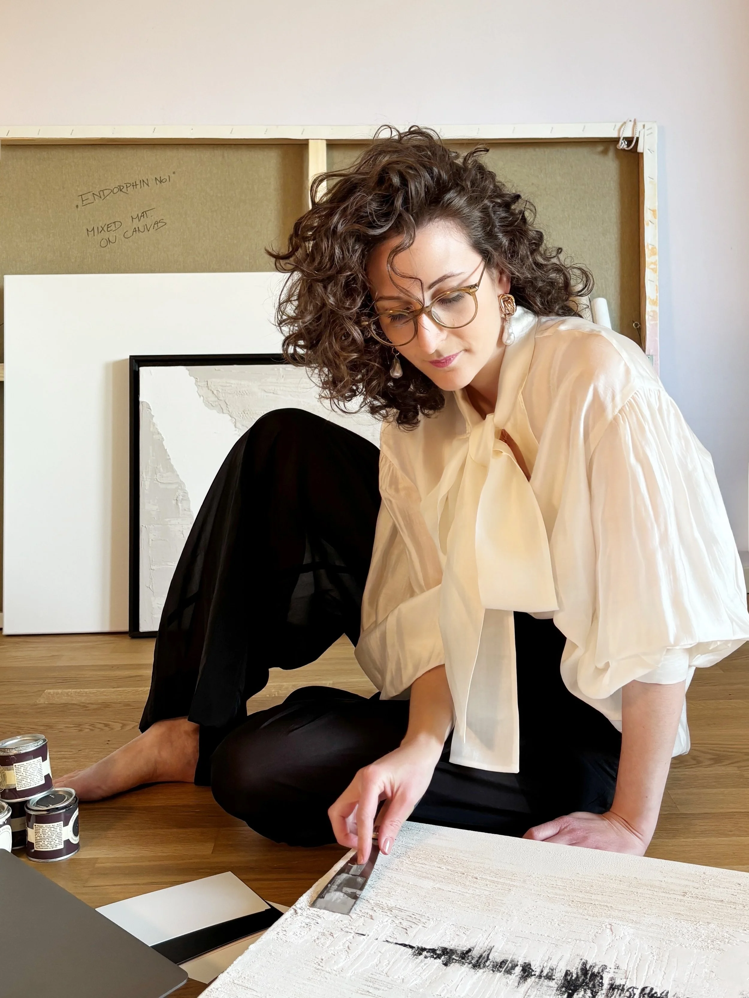 Eine Frau mit lockigem Haar, Saskia Bartels, Brille und goldenen Ohrringen arbeitet auf dem Boden an einer Kunstseite, während sie Pinsel und Farben benutzt. Im Hintergrund befinden sich Kunstwerke und eine Pinnwand.