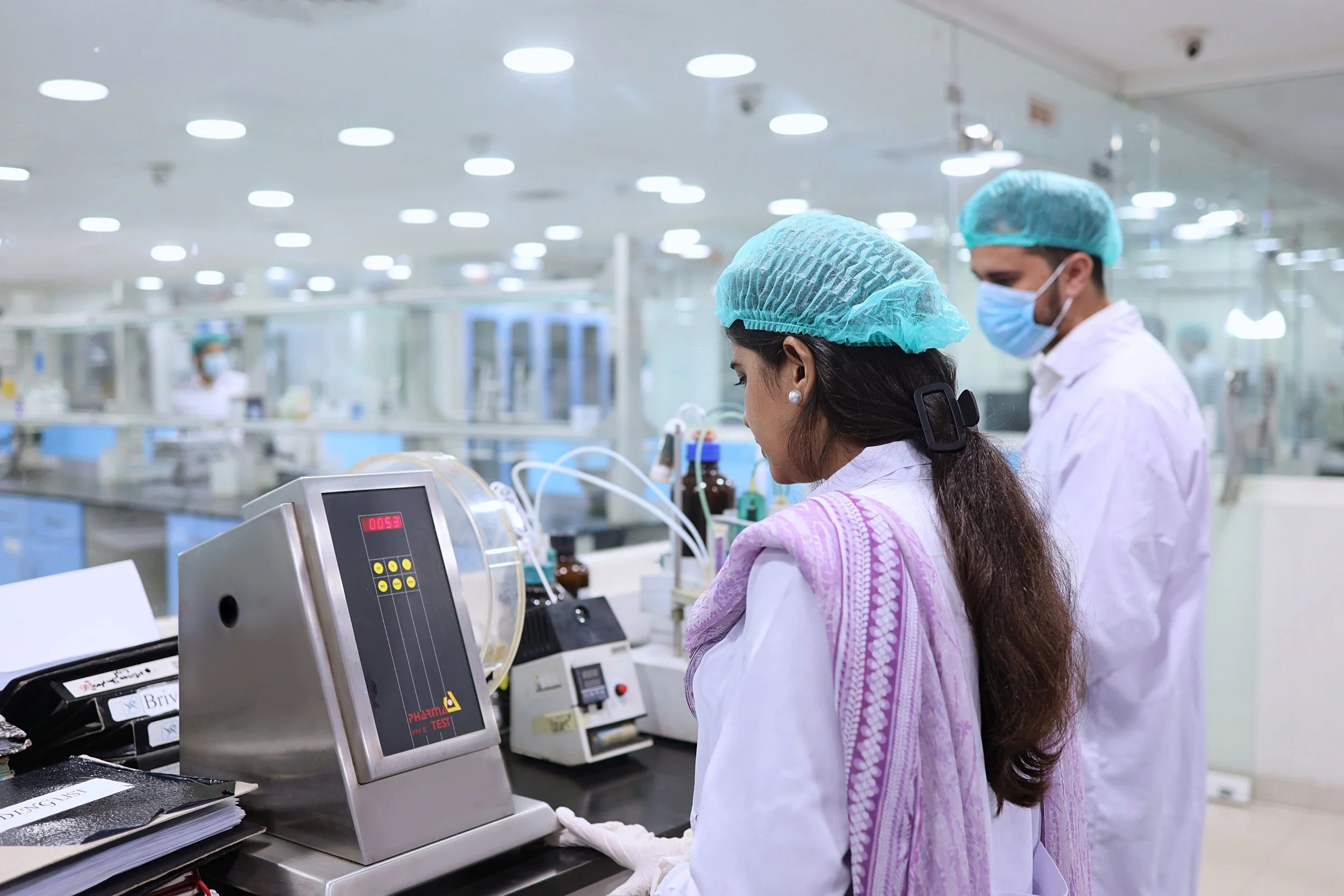 Two healthcare professionals wearing blue surgical caps, masks, and white coats working in a laboratory or hospital setting with various scientific equipment and papers on the table.