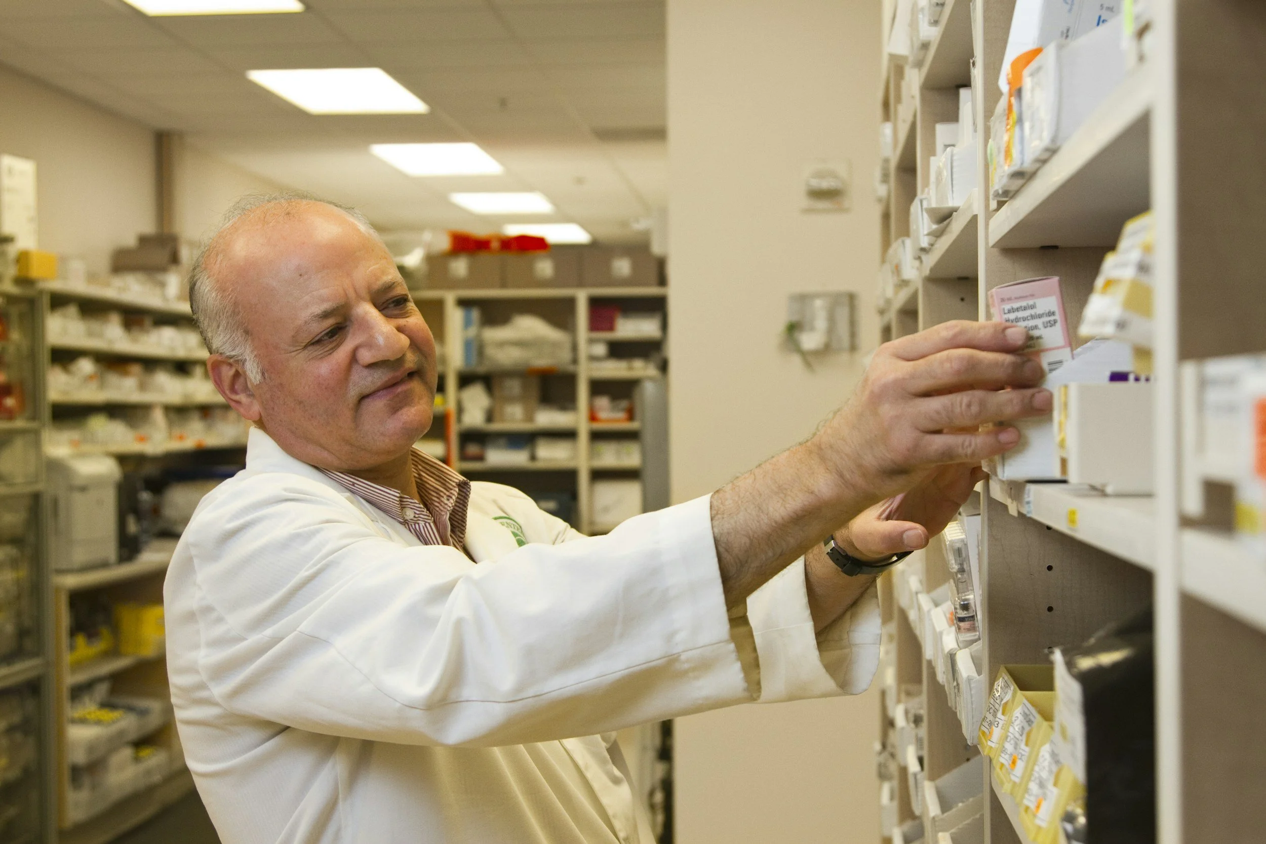 A man in a white lab coat is organizing medication or supplies on a shelf in a pharmacy or medical storage room.
