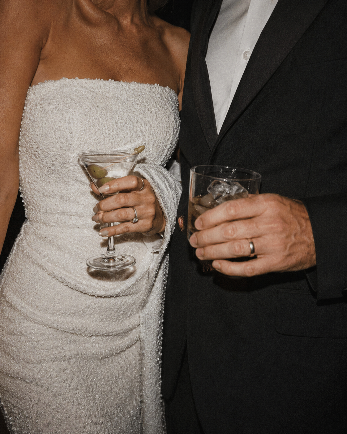 A close-up of a bride and groom holding drinks at their wedding. The bride is wearing a strapless, textured white dress, and the groom is dressed in a black suit with a white shirt. Both are wearing wedding rings.