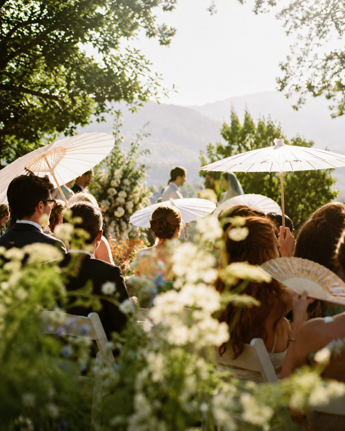 Outdoor wedding ceremony with guests sitting in chairs, some holding white parasols, surrounded by trees and flowers with mountains in the background.