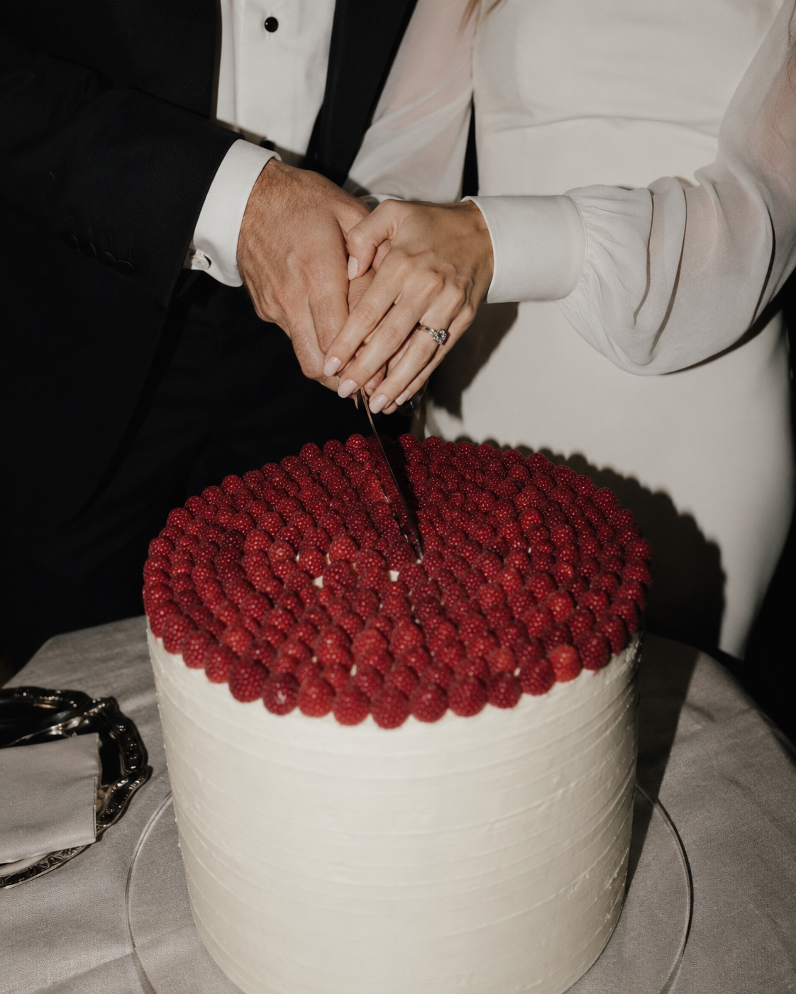 A couple's hands holding a knife together, preparing to cut a round wedding cake topped with raspberries.