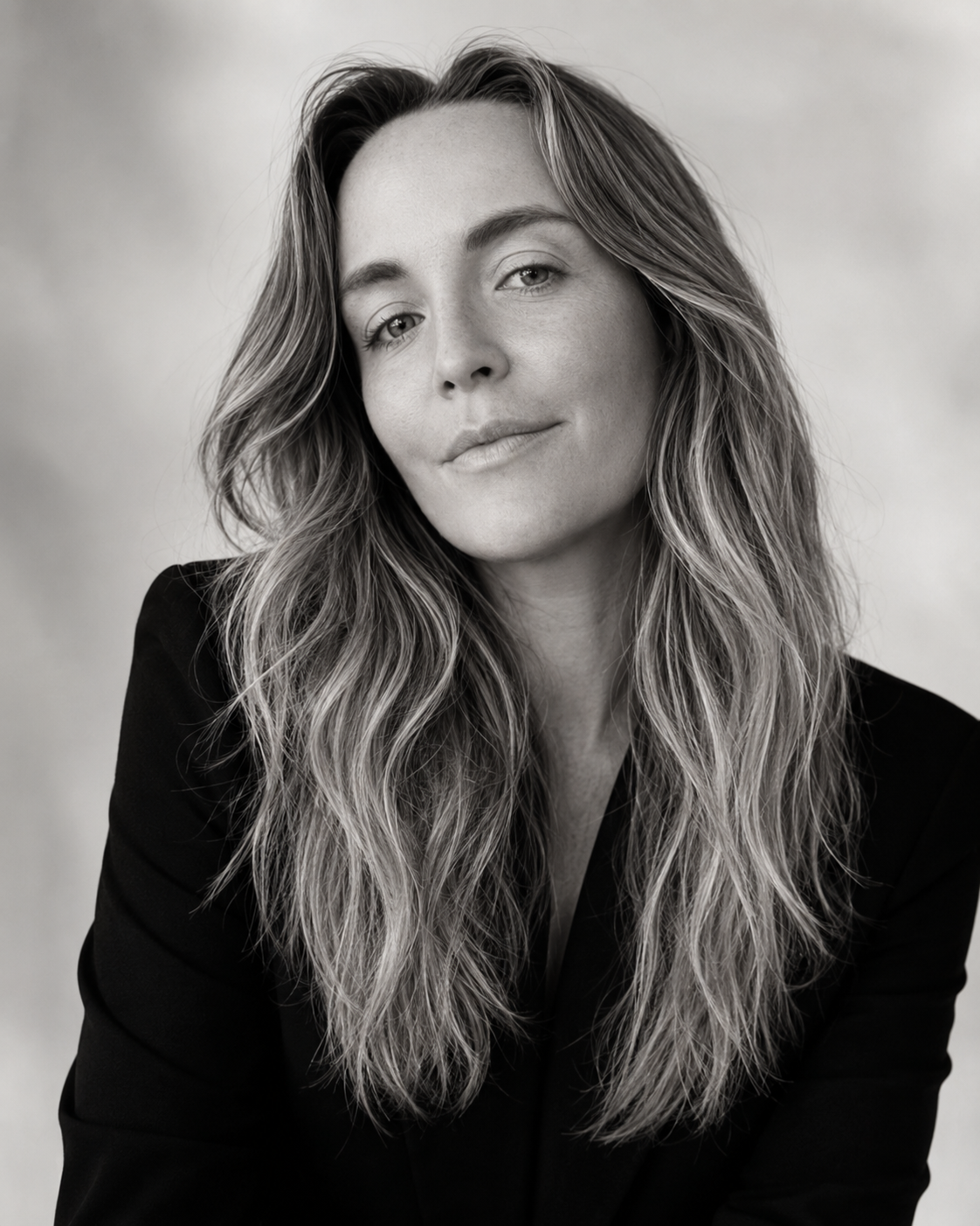 Black and white portrait of a woman with long wavy hair, wearing a dark blazer, looking at the camera with a slight smile.