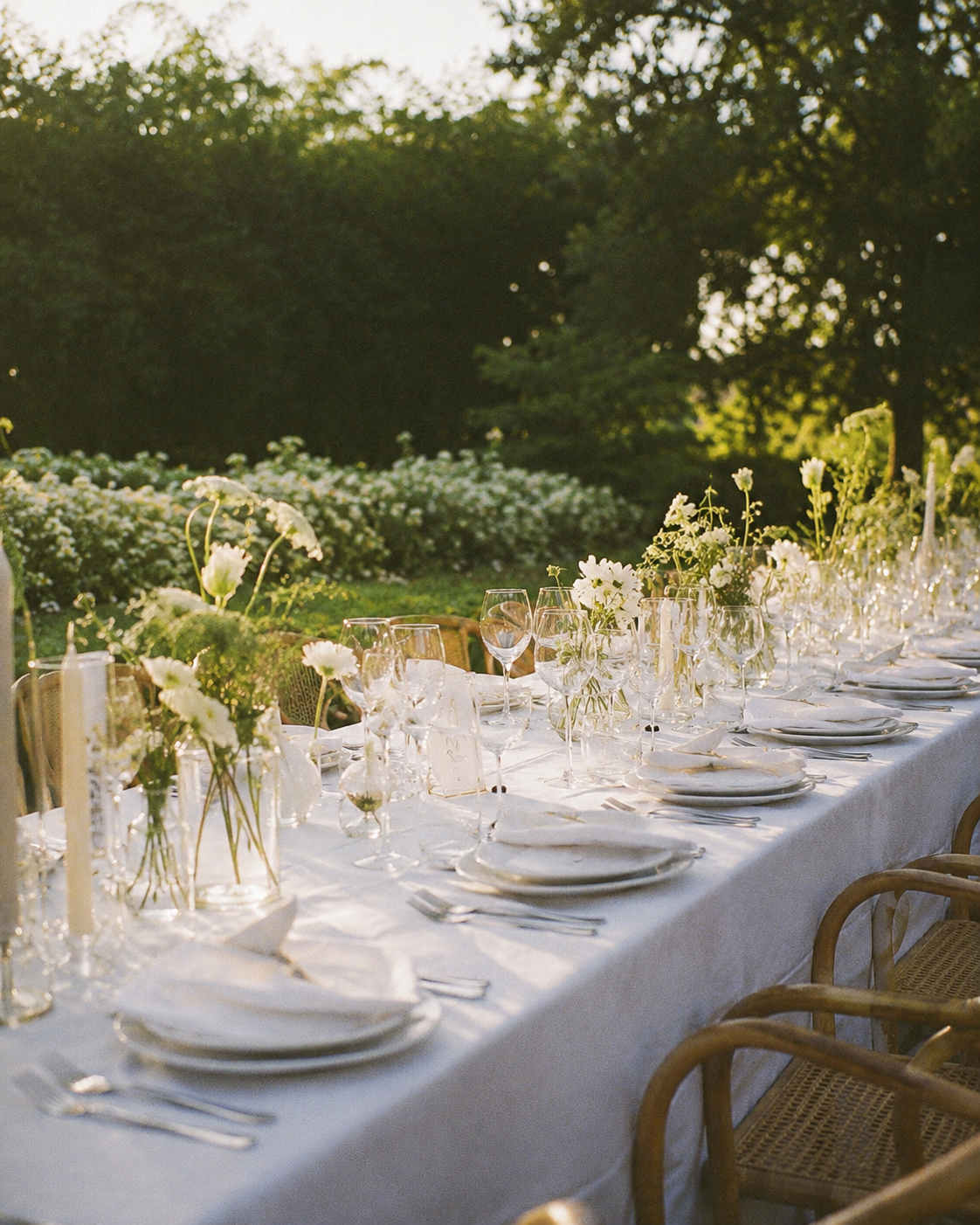 A long outdoor dining table set for a formal event, decorated with white flowers and surrounded by wicker chairs, on a sunny day in a garden.