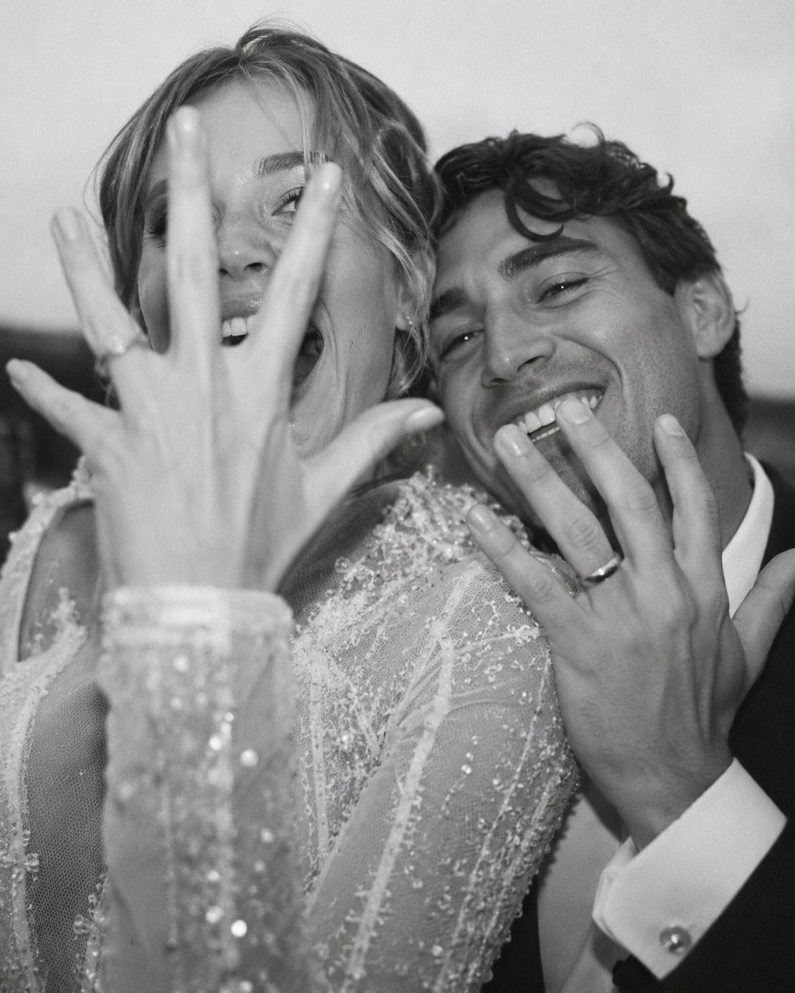 A black and white photo of a joyful couple, presumably at a wedding, with the woman wearing a beaded dress and the man in a tuxedo. They are smiling and playfully touching each other's faces.
