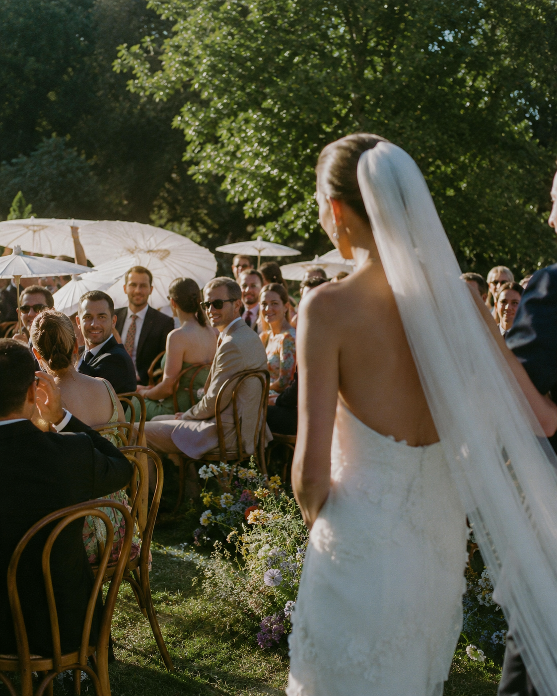 A bride with long blonde hair and a white wedding dress stands outdoors during a wedding ceremony, surrounded by seated guests holding umbrellas in the bright sunlight.