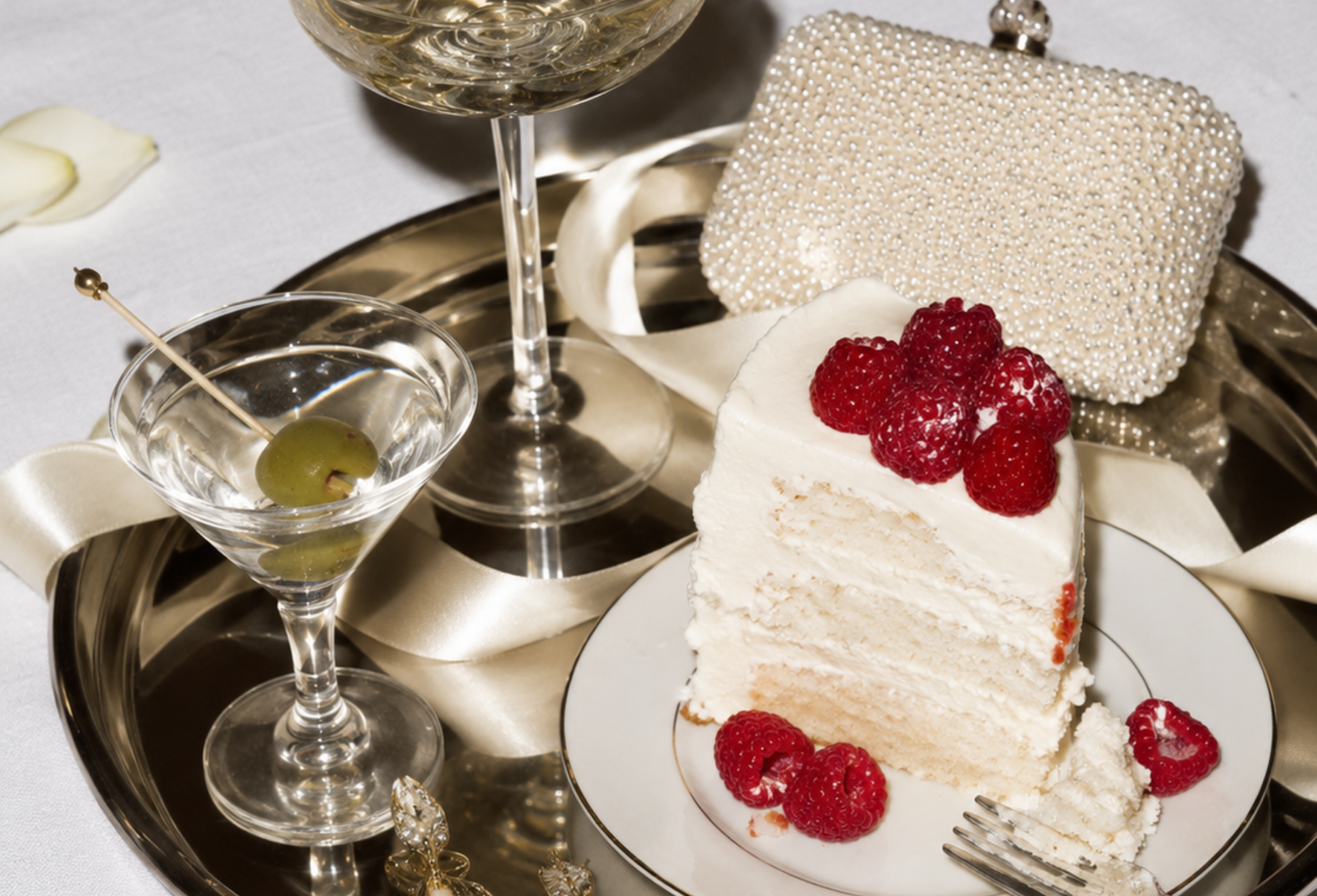 A slice of white cake with raspberries on top, a glass of white wine, a cocktail with an olive, and a decorative clutch purse all arranged on a silver tray.