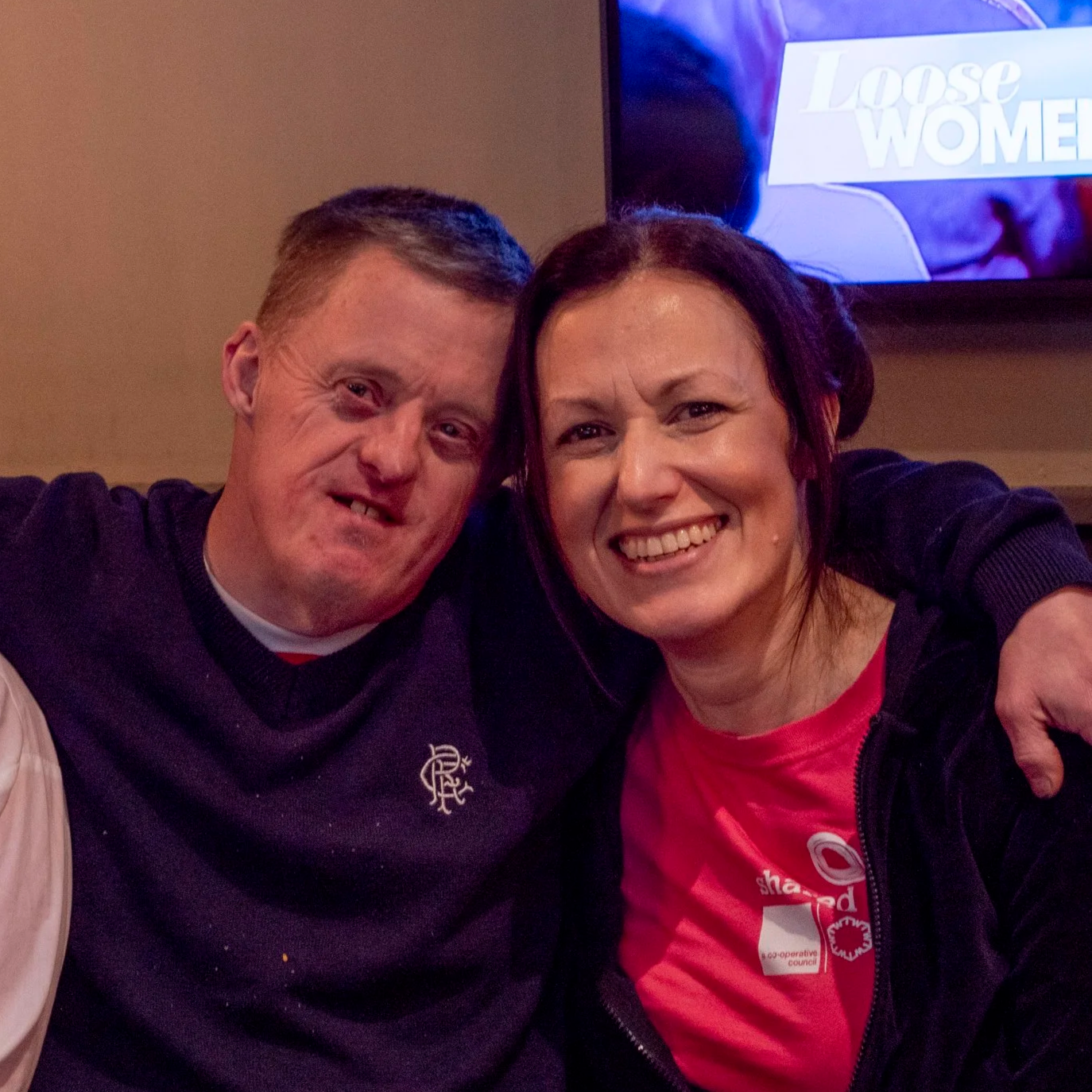 Two smiling people, a man and a woman, sitting closely together with their arms around each other, in a casual indoor setting.