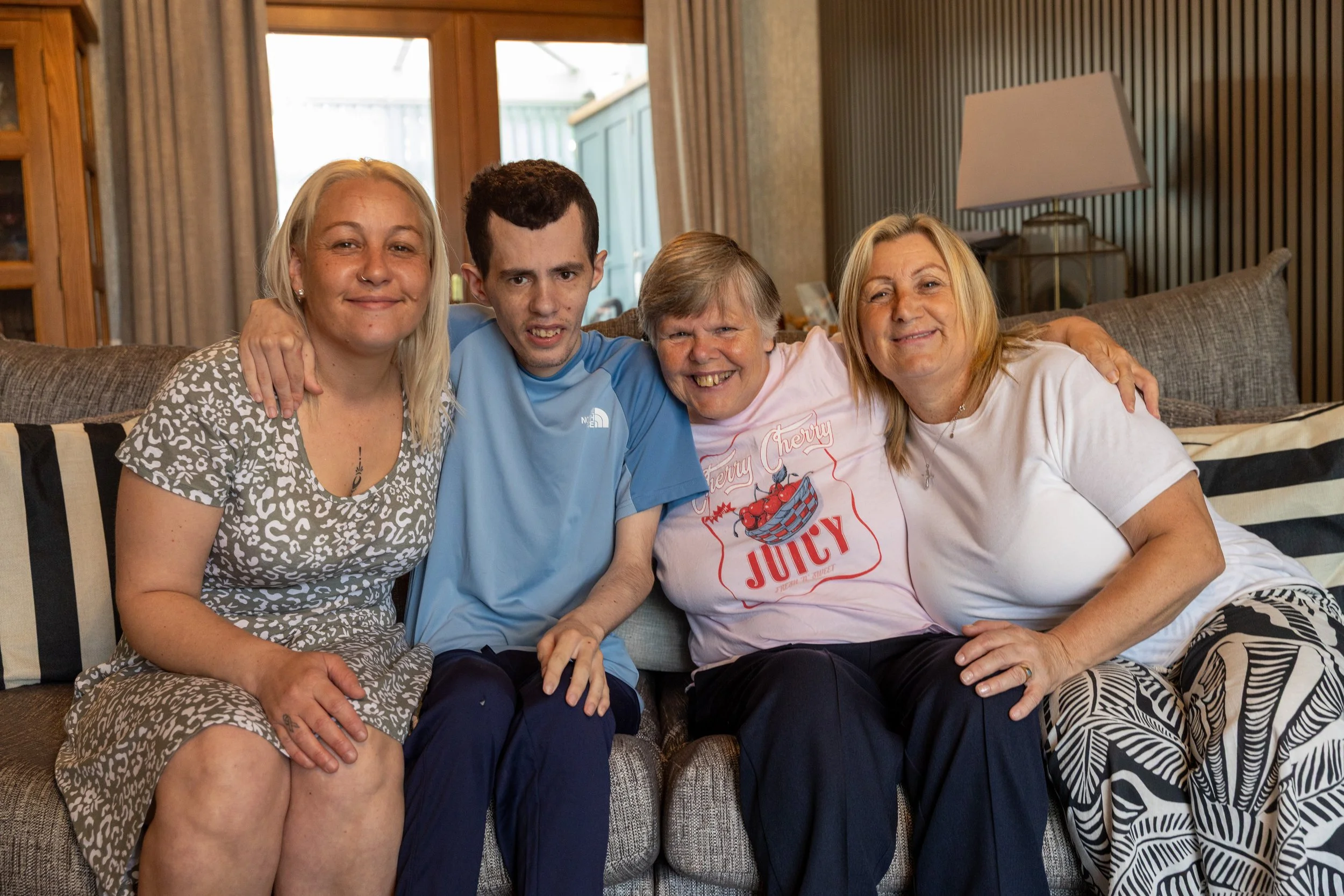 Four people sitting on a sofa in a living room, smiling, with two women on each side of two men, embracing each other.