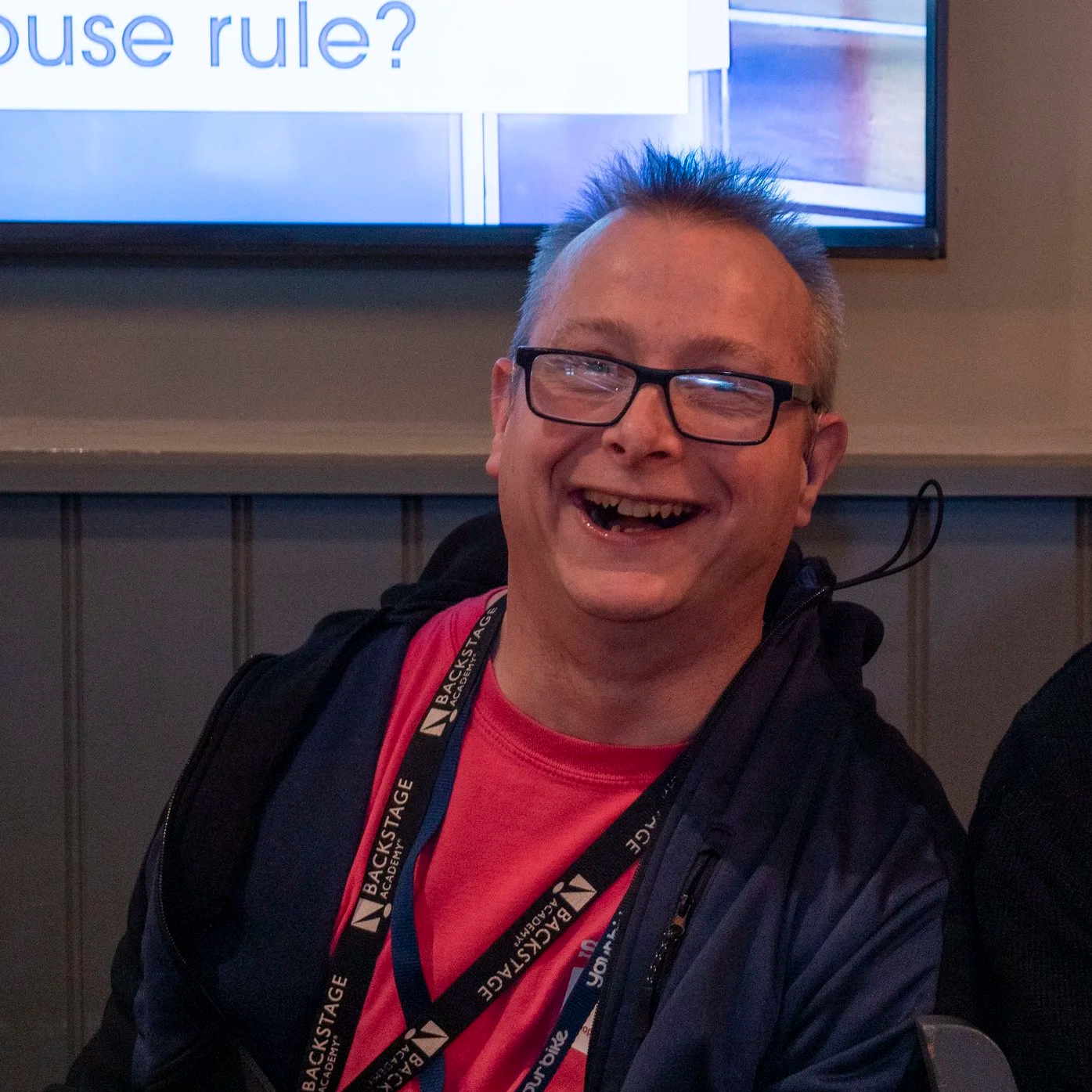 A smiling man with glasses, wearing a red shirt and black jacket, sitting indoors in front of a television screen displaying text. He has a lanyard around his neck.