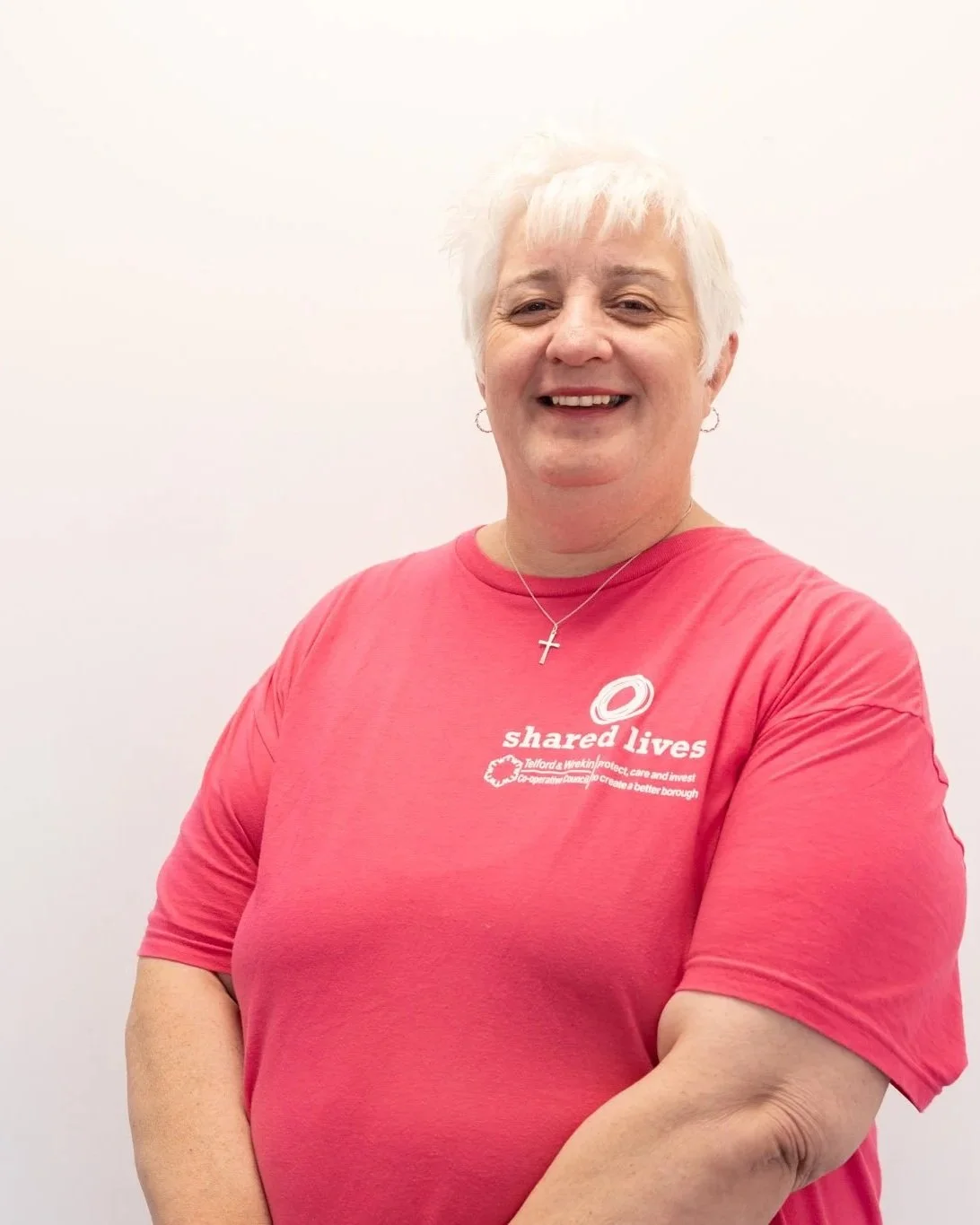Smiling woman with short white hair wearing a pink t-shirt with shared lives logo, silver cross necklace, and hoop earrings, standing against a plain white background.