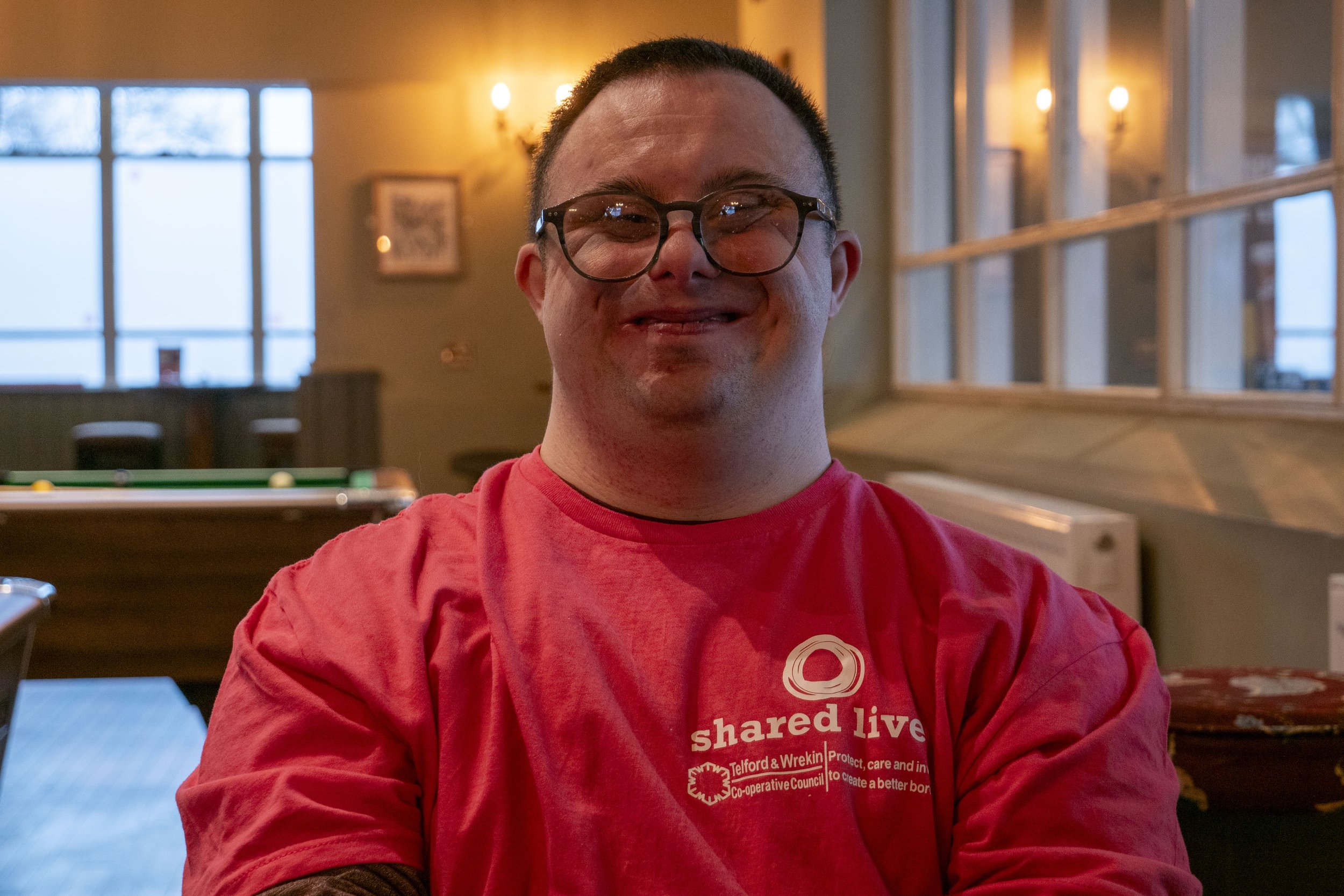 A man with short dark hair, glasses, and a goatee smiling in a cozy indoor setting with warm lighting, wooden accents, and large windows in the background.