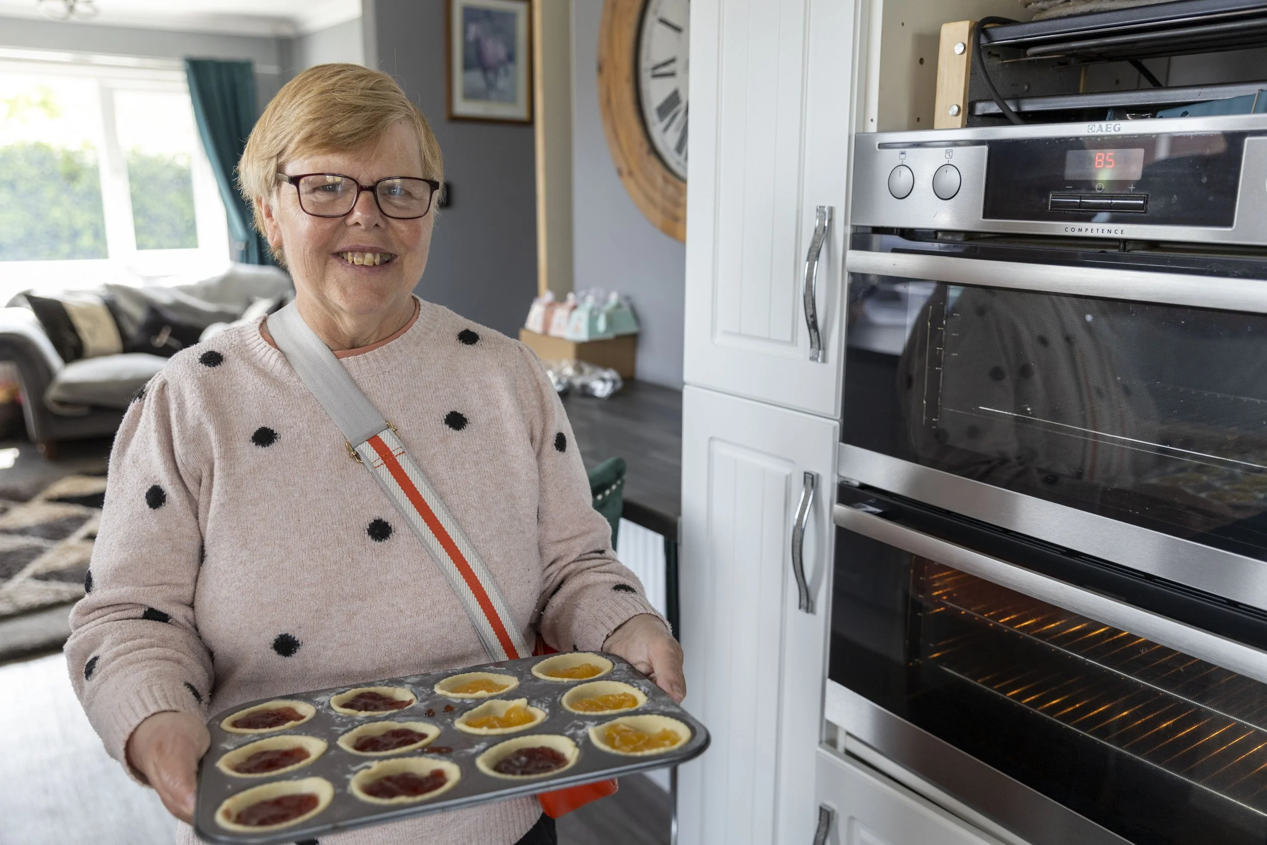 An elderly woman wearing glasses and a pink sweater with black polka dots, holding a tray of mini pies with various fruit toppings, standing in a kitchen near an oven.