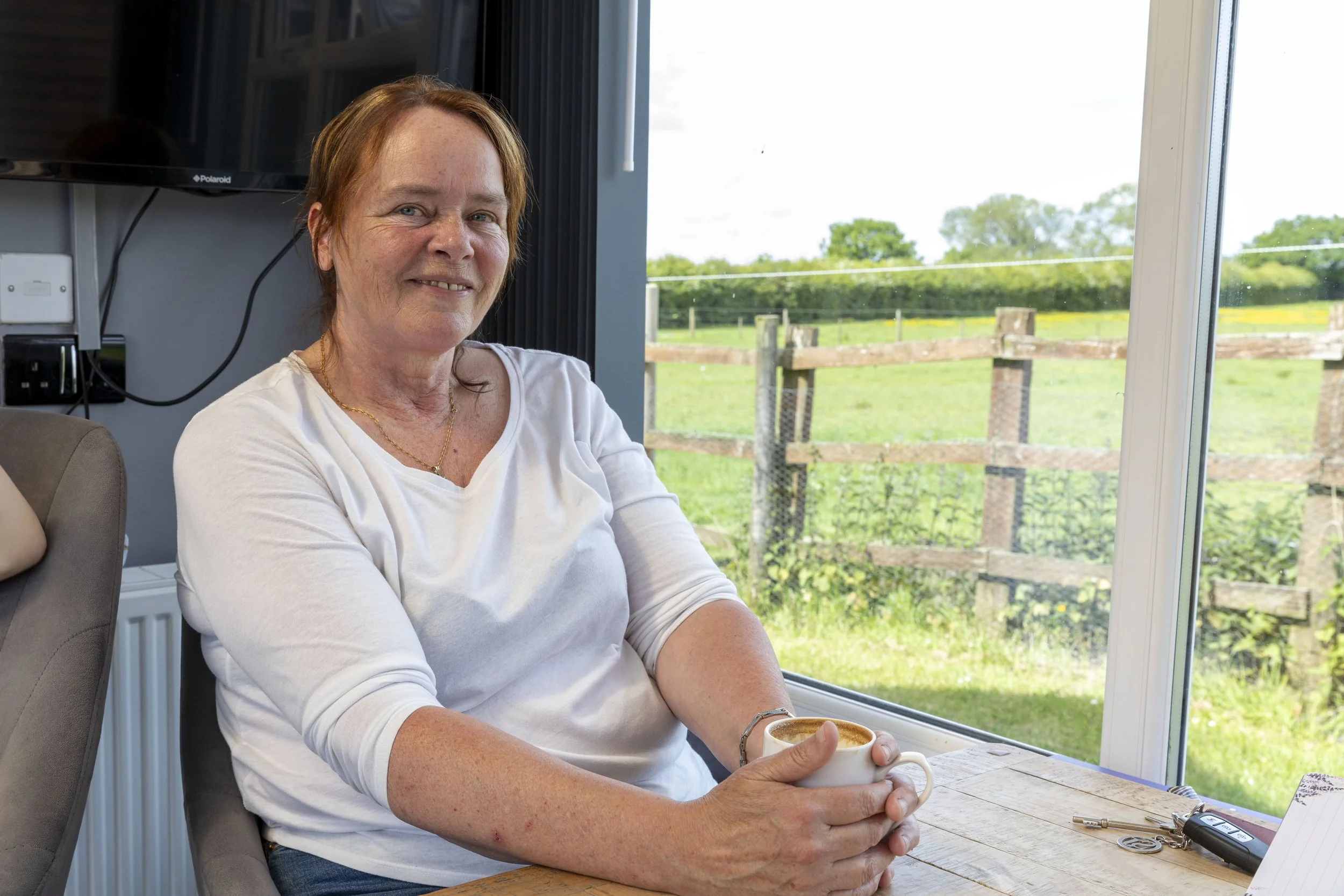 A woman with red hair and a white shirt sitting at a wooden table, holding a cup of coffee, near a large window with a scenic outdoor view of green fields and trees.