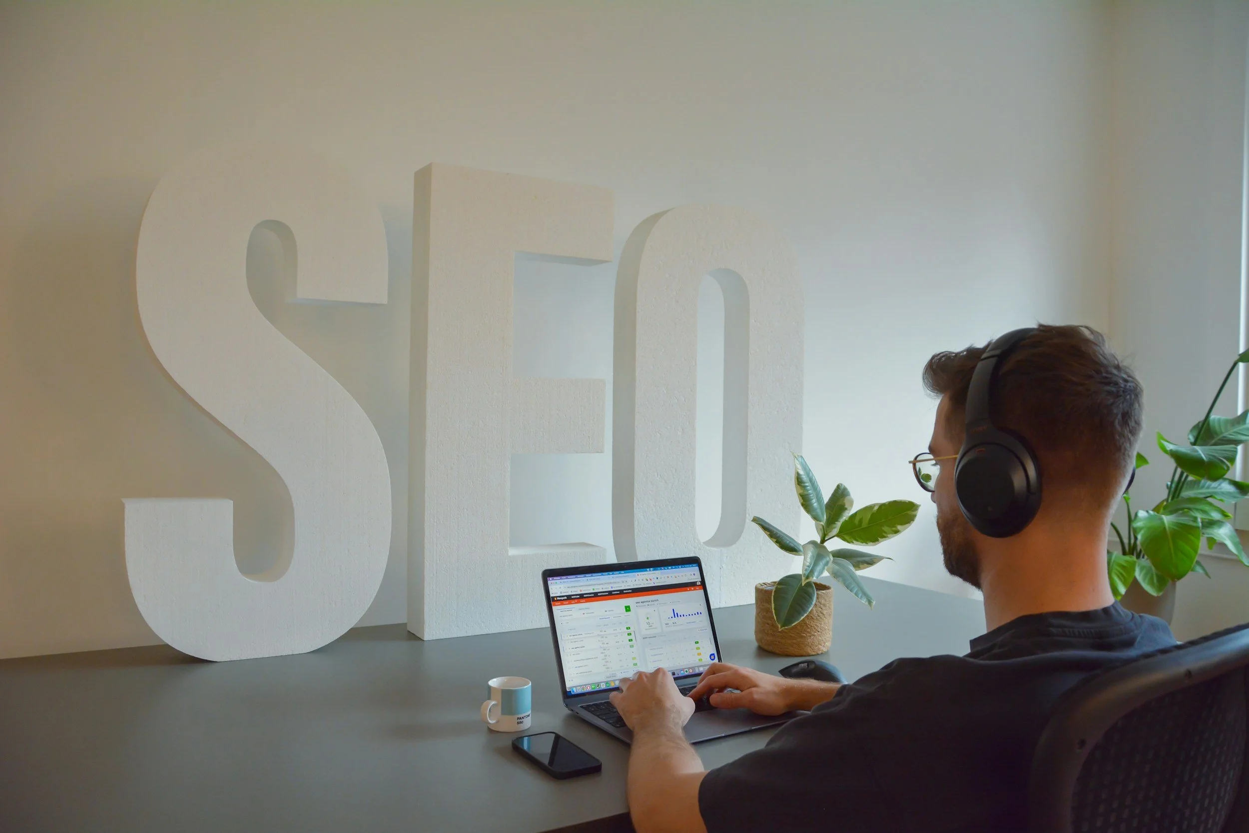 A man with brown hair wearing glasses and large headphones working on a laptop at his desk in an office. There are plants on the desk and a large white decorative letter 'S' on the wall behind him.