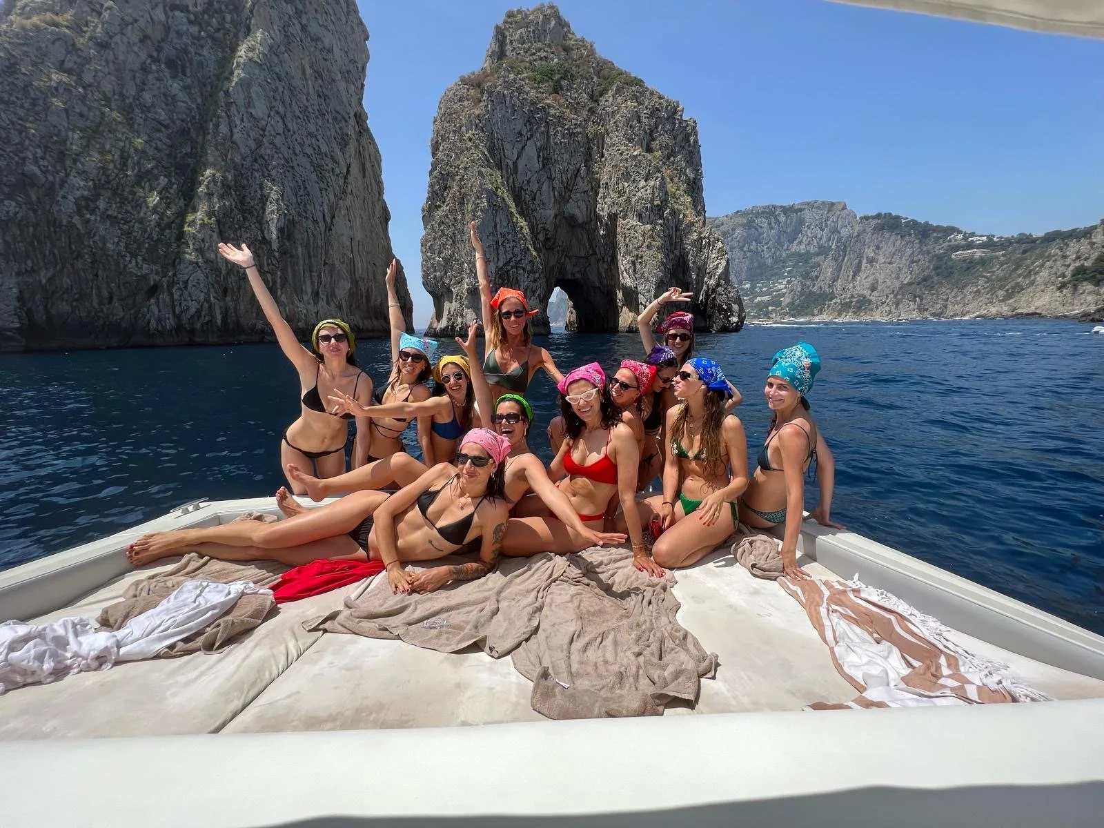 Group of women in swimsuits with colorful headscarves and sunglasses enjoying a boat trip in a scenic coastal area with large rock formations, clear blue water, and mountainous landscape in the background.