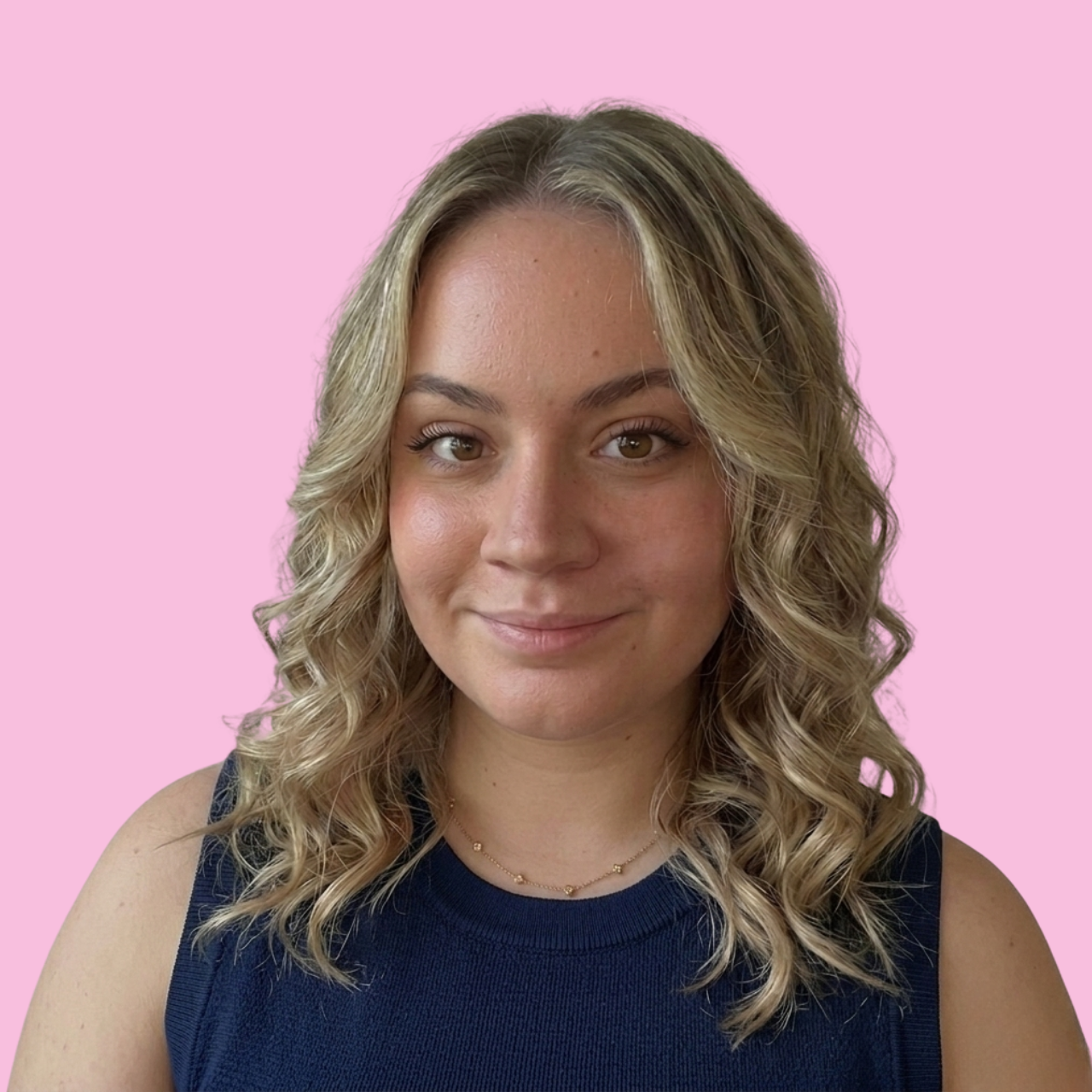 Portrait d'une femme souriante avec des cheveux bouclés blonds, portant un haut sans manches bleu foncé, contre un fond rose clair.