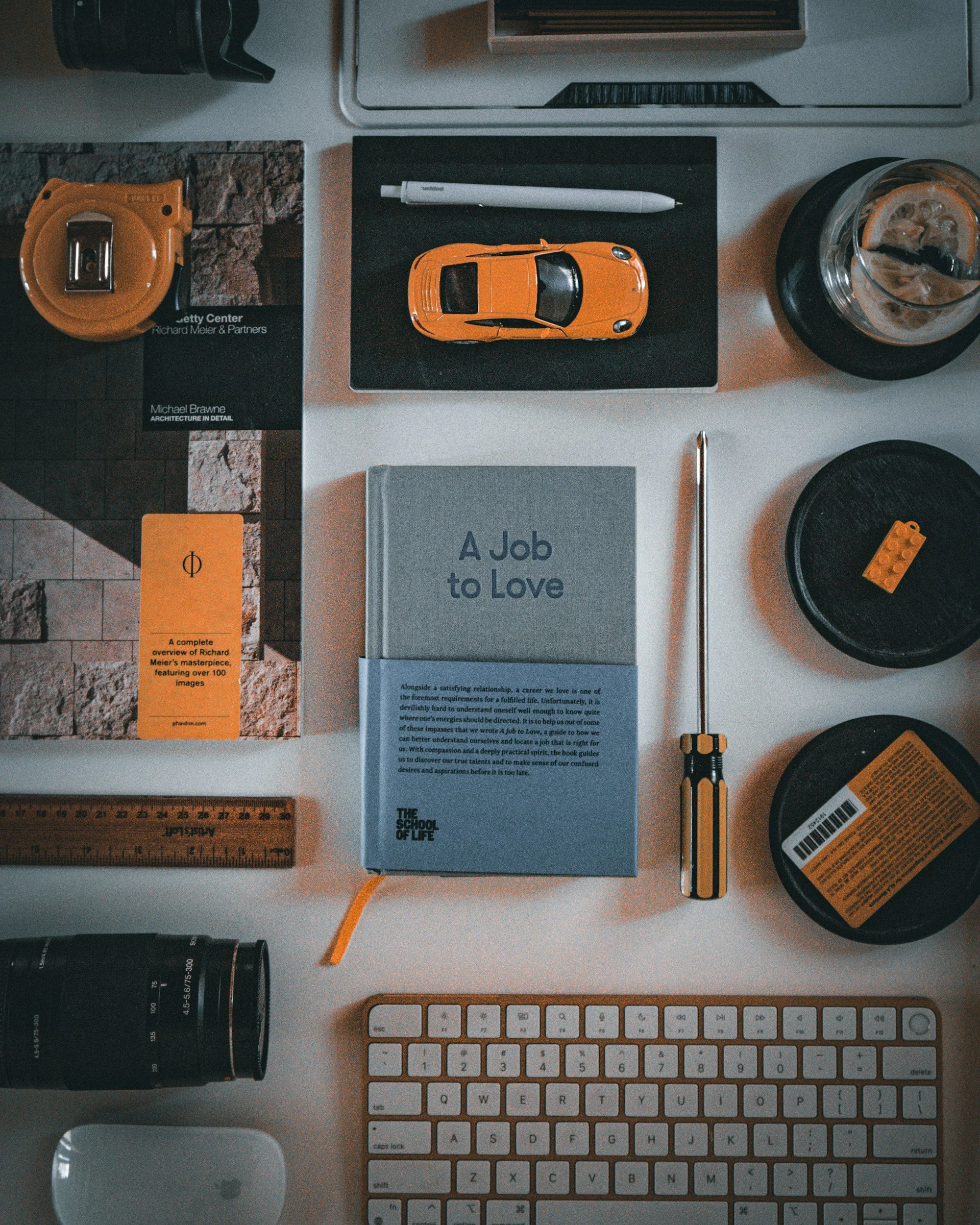 A desk scene with various items including a toy yellow sports car, a white pen, a book titled 'A Job to Love', a small notebook, a glass of iced coffee, a yellow screwdriver, a tape measure, a ruler, a camera lens, a pair of black headphones, an Apple Magic Mouse, and a white keyboard. The background shows a brick wall and a computer monitor.