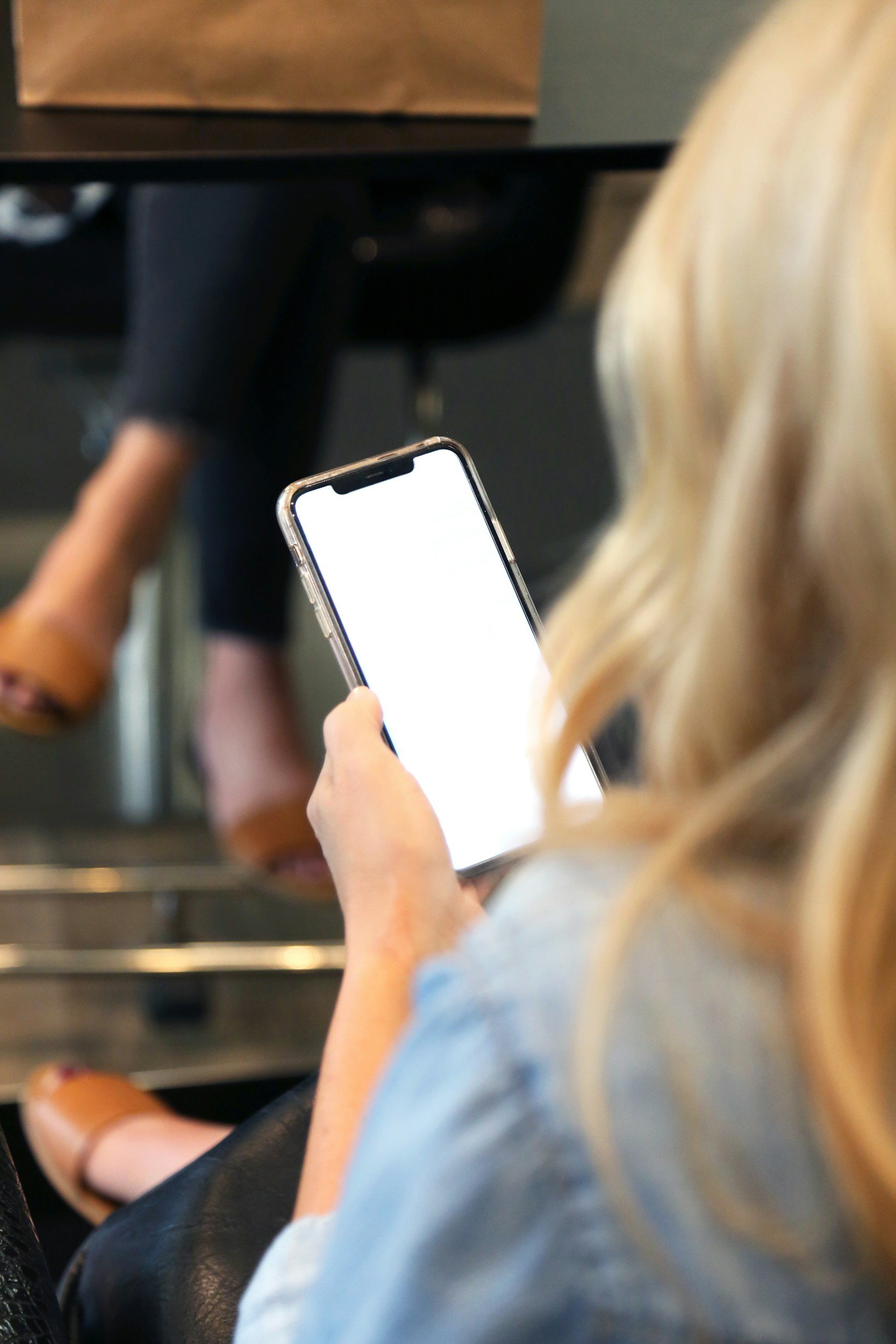 A person with blonde hair wearing a light blue shirt sitting on a black leather seat, holding a smartphone in their left hand. Part of their right arm and a brown sandal on their foot are visible. The person seems to be in a public space, like a train or bus, with a dark floor and a table or counter in front of them.