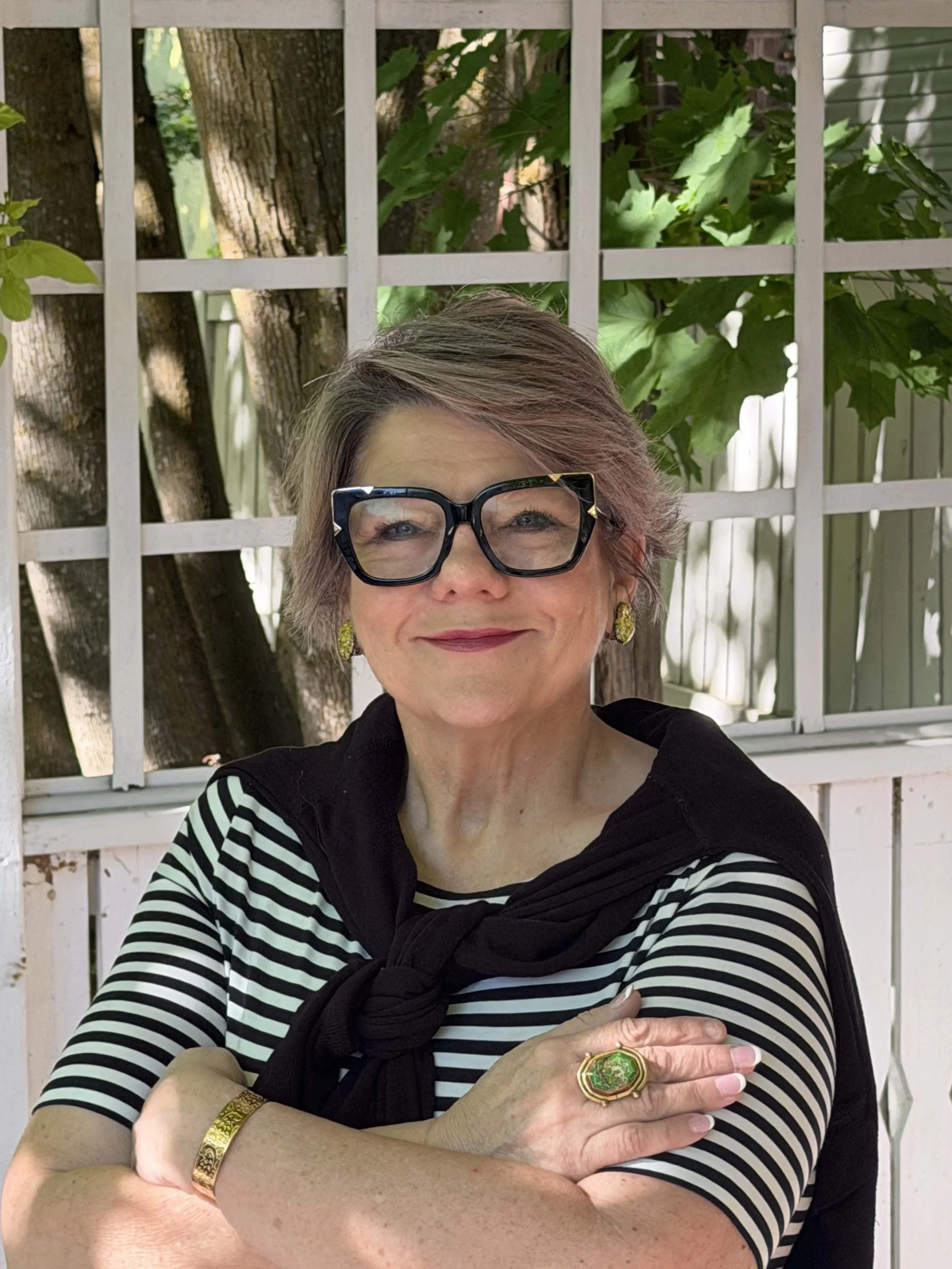 Portrait of Krys, founder of The Accessory Alchemist, wearing signature glasses and vintage jewelry while seated outdoors in a garden setting.