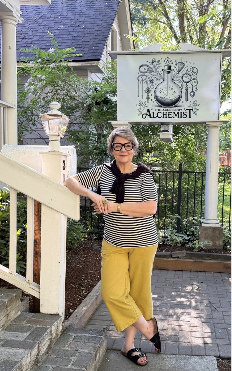 Founder of The Accessory Alchemist standing outside the boutique beneath the brand sign featuring the signature alchemy bottle logo.