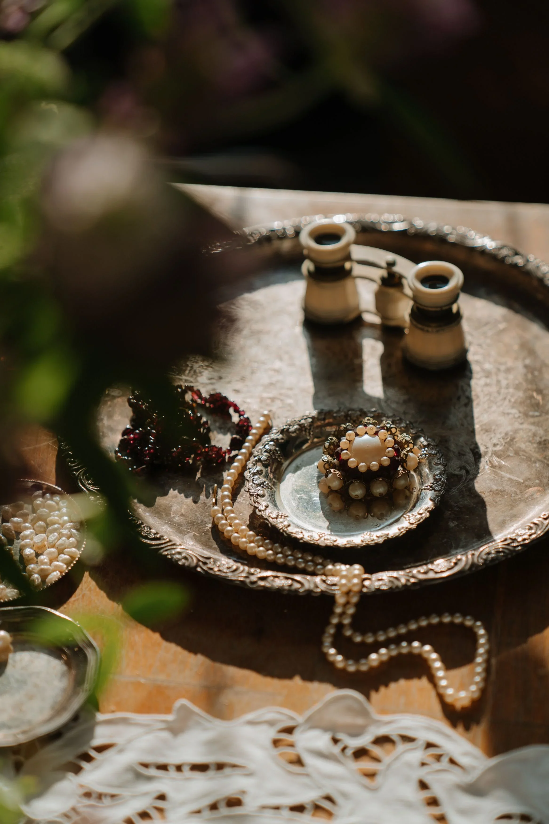 Vintage pearl necklace and jeweled brooch arranged on an antique silver tray in warm natural light.