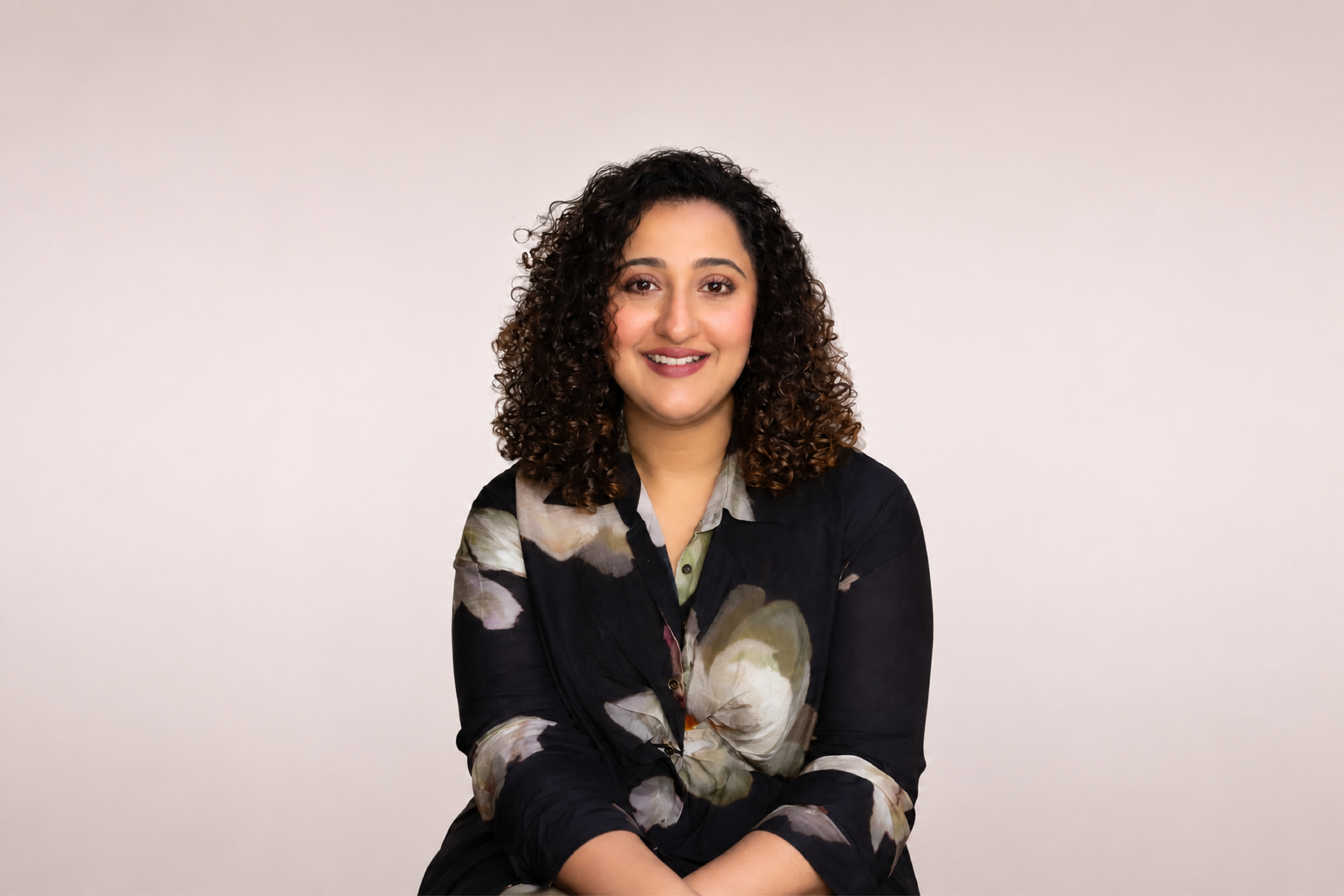 A woman with curly brown hair smiling and sitting against a plain light-colored wall. She is wearing a dark blouse with a floral pattern.