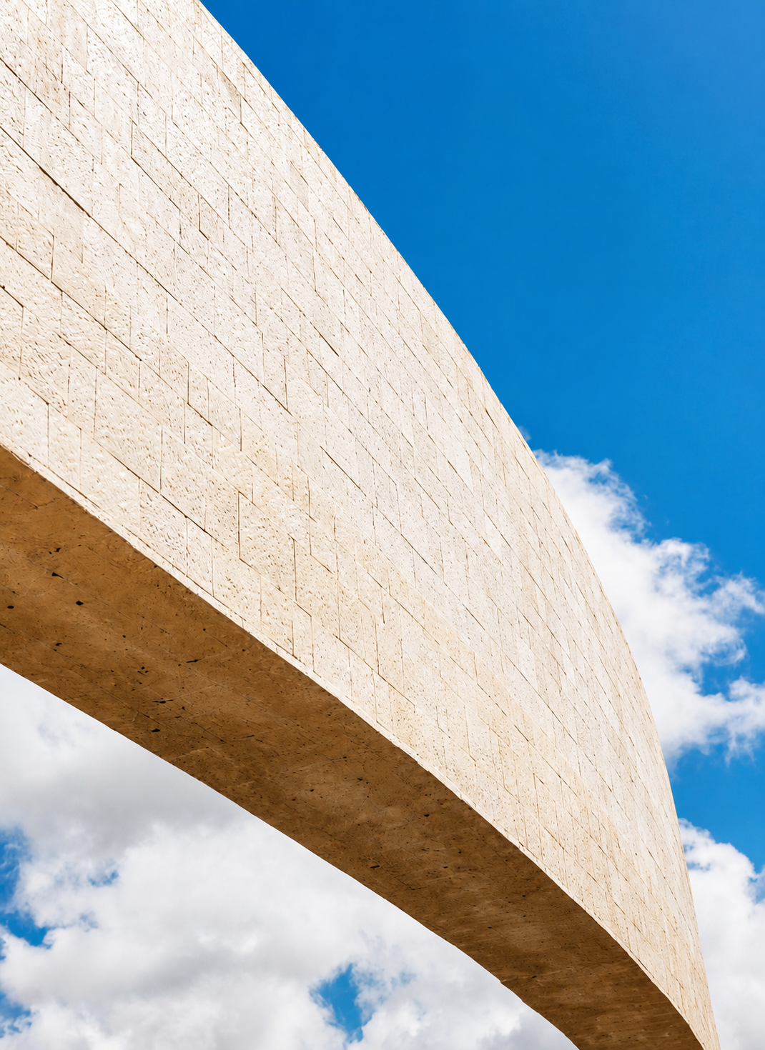 Low-angle view of a large, curved beige stone or concrete structure against a bright blue sky with some clouds.