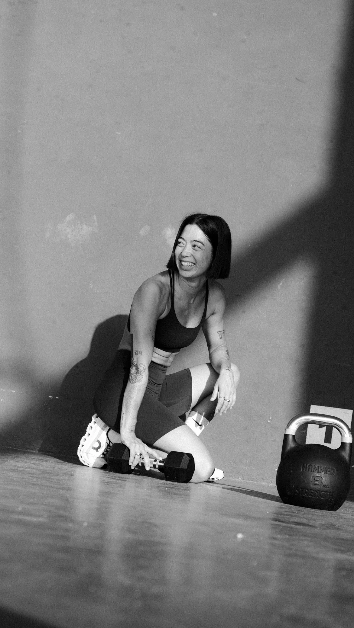 Claris the founder of the wholesome workout leaning against dumbells and kettlebells happy to everyone landing on this page and ready to help people transform.