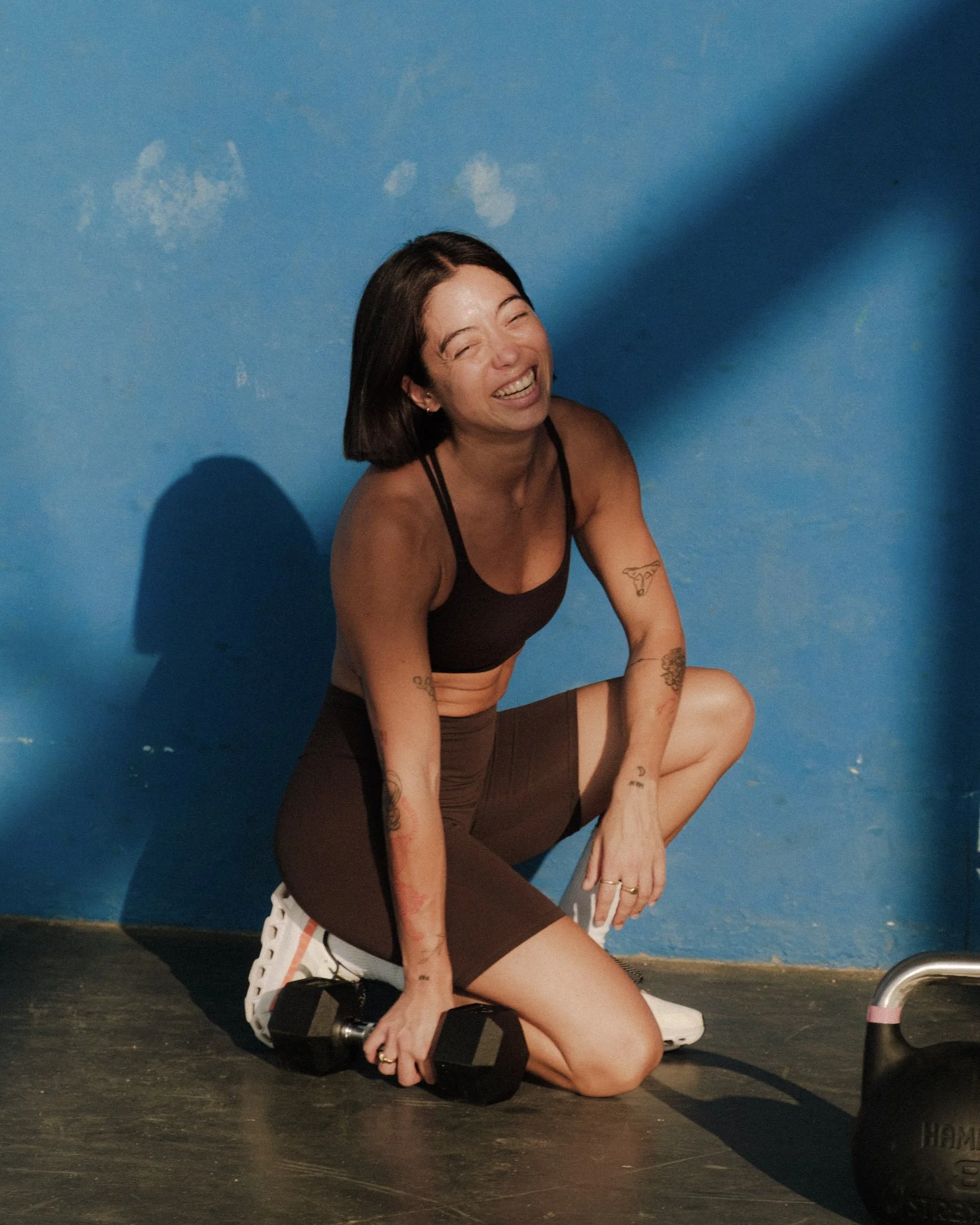 Claris the founder of the wholesome workout leaning against dumbells and kettlebells happy to everyone landing on this page and ready to help people transform.